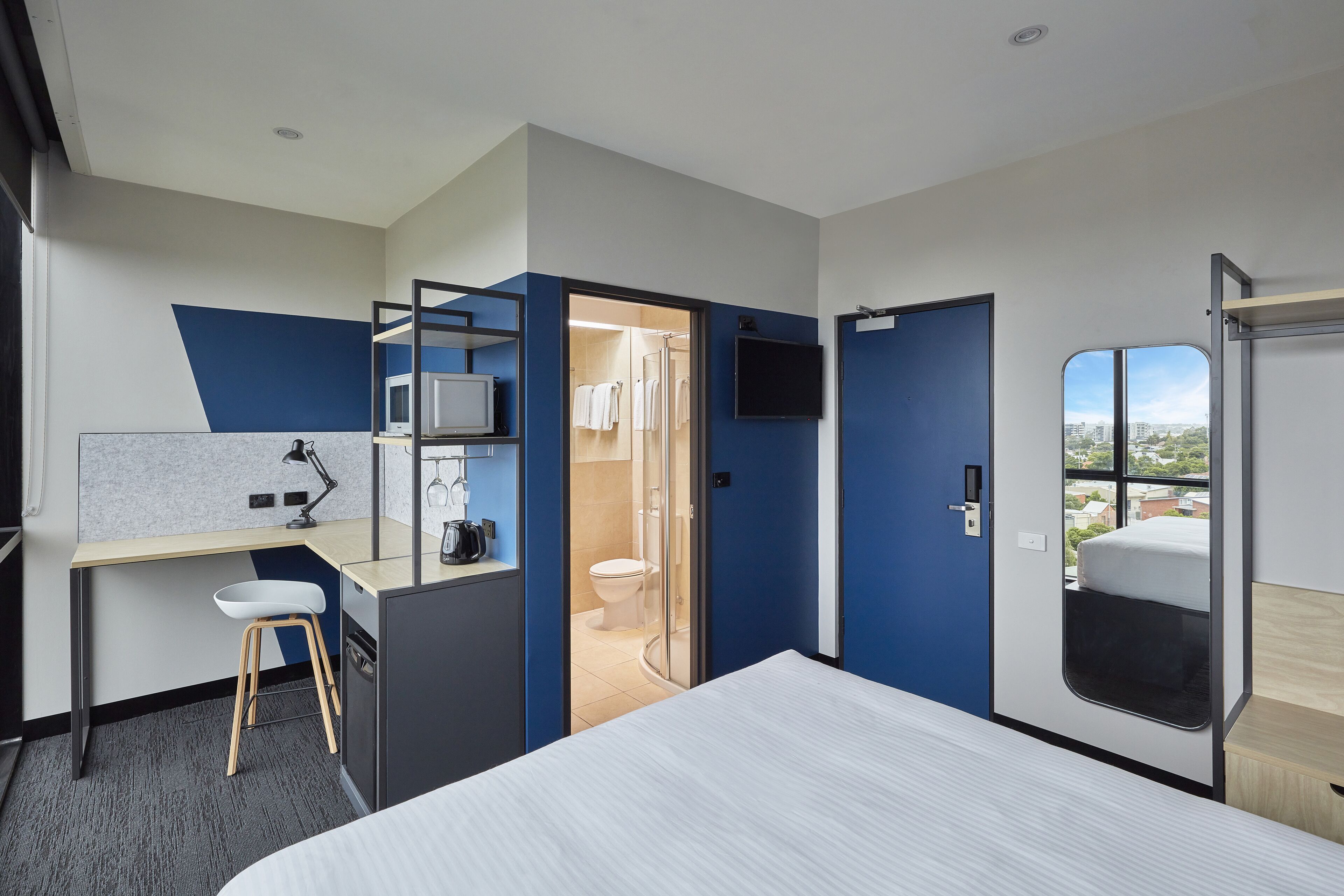 A modern hotel room with a bed, a desk with a chair, a TV, and a view of the city through a window.