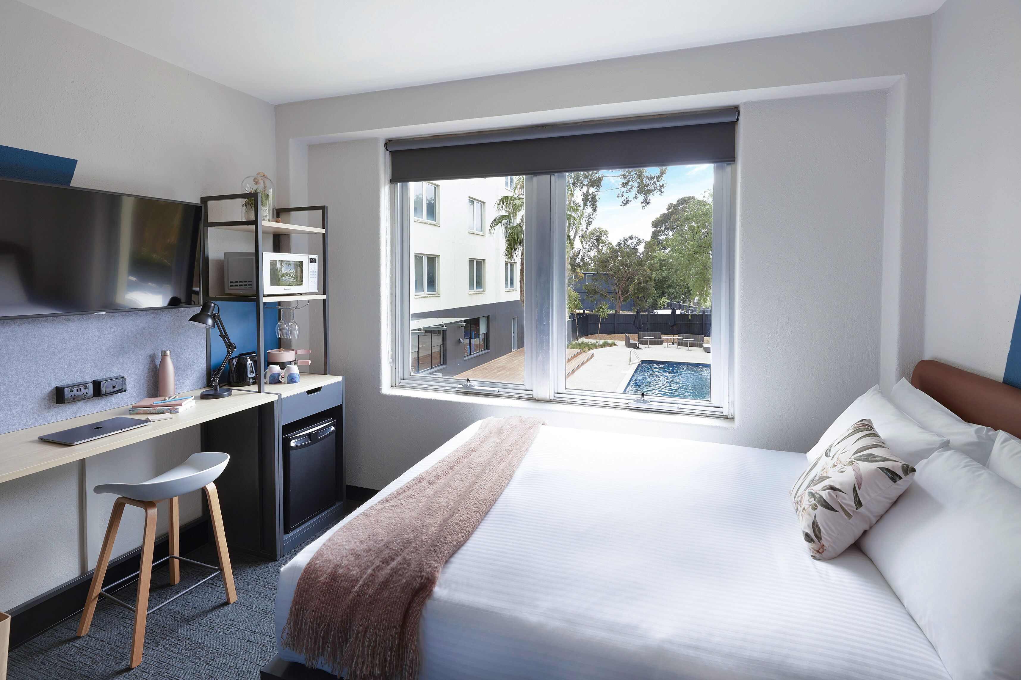 A modern hotel room with a bed, a desk, a chair, a TV, a microwave, and a window with a pool view.