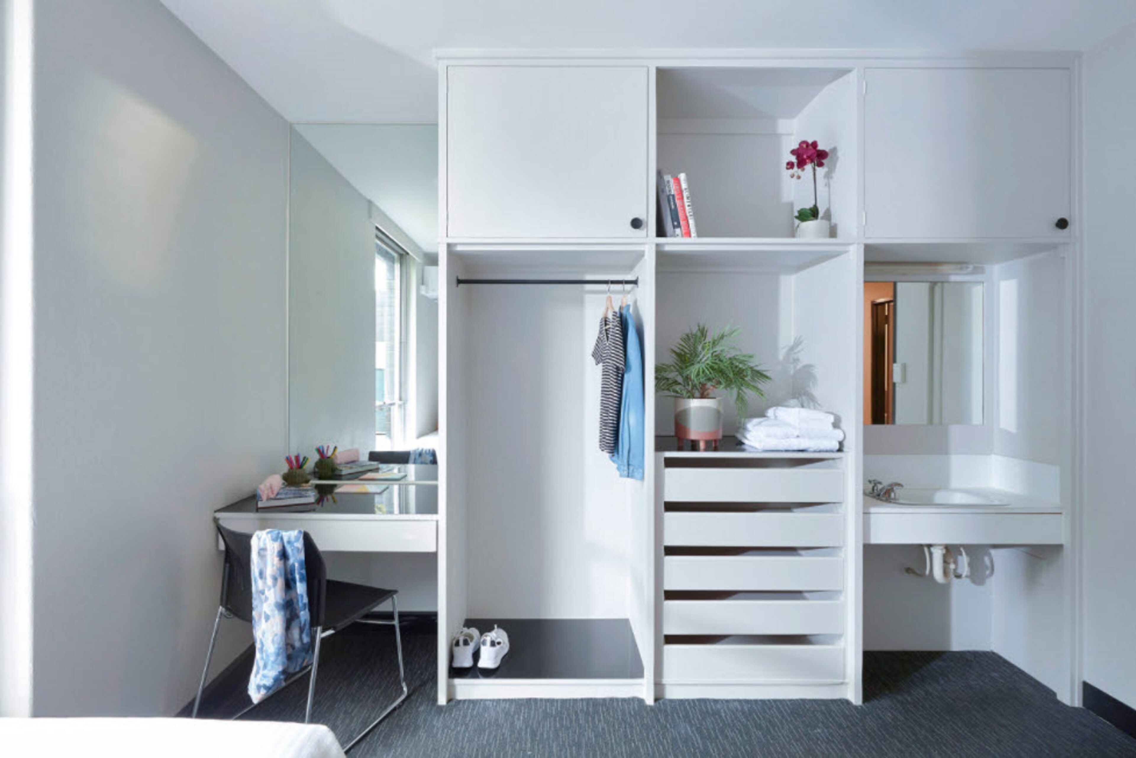 Habitación de hotel con escritorio, armario, espejo, lavamanos y una planta en maceta.