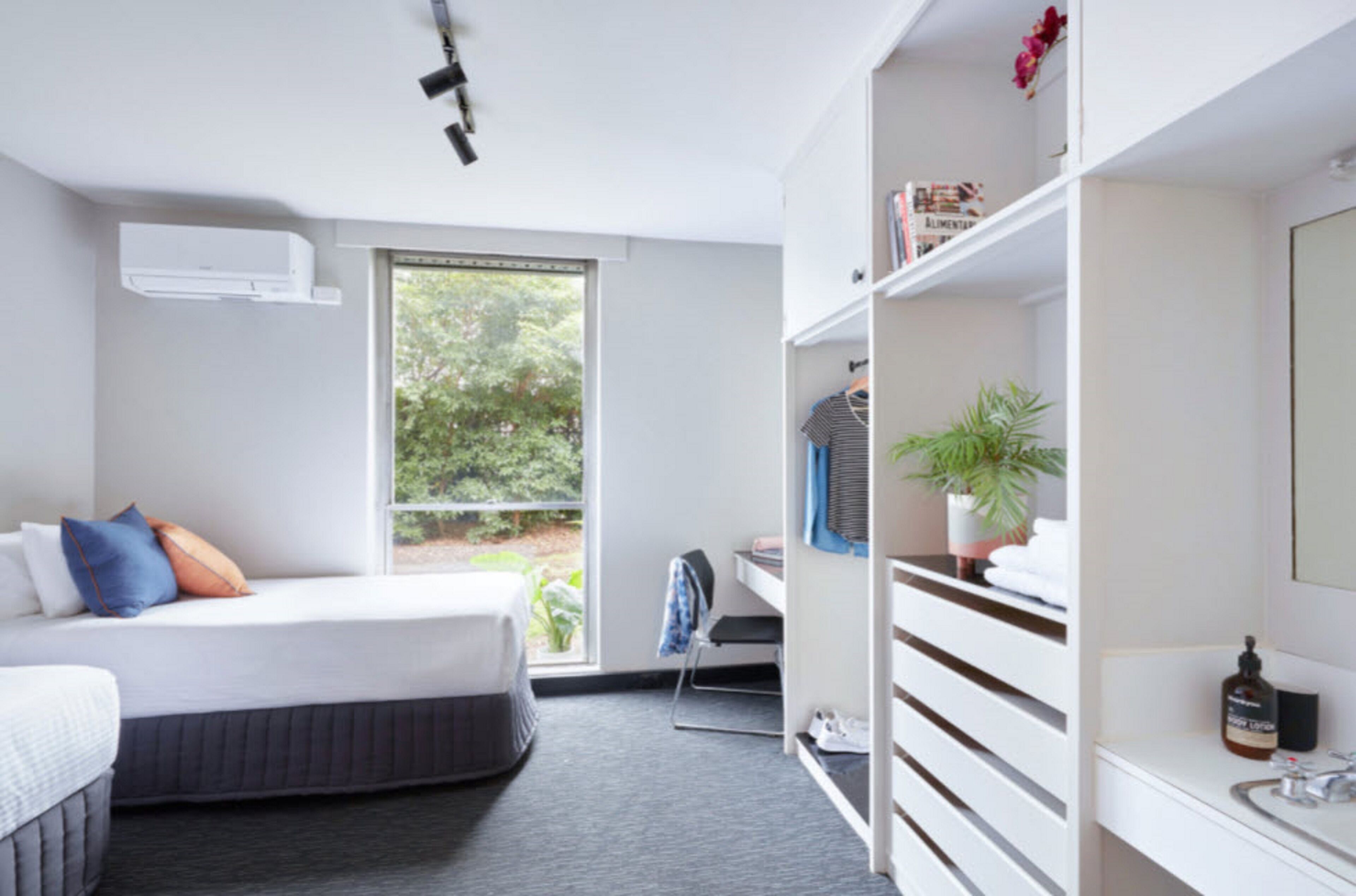 Quarto de hotel moderno com cama, escrivaninha, cadeira, guarda-roupa e janela com vista para o verde.