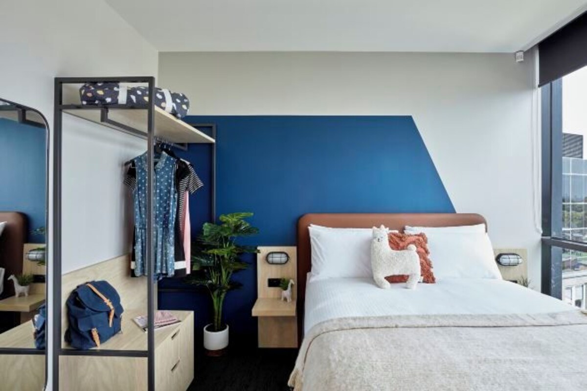 A modern hotel room with a bunk bed, a desk with a backpack, a potted plant, and a large window.