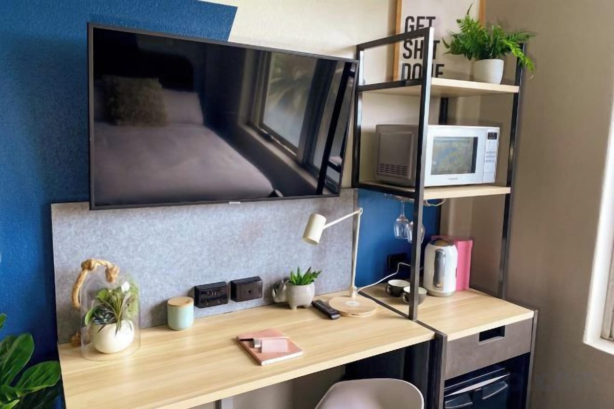 A modern room with a flat-screen TV mounted on the wall, a desk with a computer, a microwave, and a shelf with plants and a coffee maker.