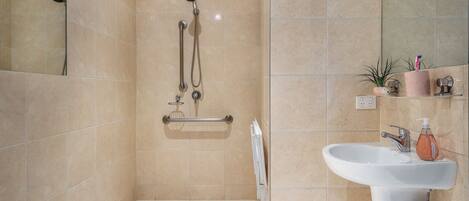 A bathroom with beige tiles, a shower with a handheld showerhead, a grab bar, a white sink with a soap dispenser, and a mirror.