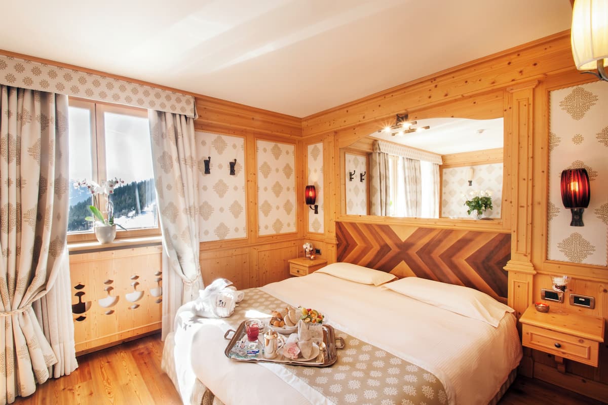 Una camera d'hotel con testiera in legno, un letto grande con un vassoio con prodotti per la colazione, una finestra con tende e uno specchio che riflette la stanza.