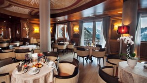 A luxurious dining area with high ceilings, ornate columns, and large windows offering mountain views. Tables are set with fine china, silverware, and floral centerpieces. - Cristal Palace (Pinzolo)