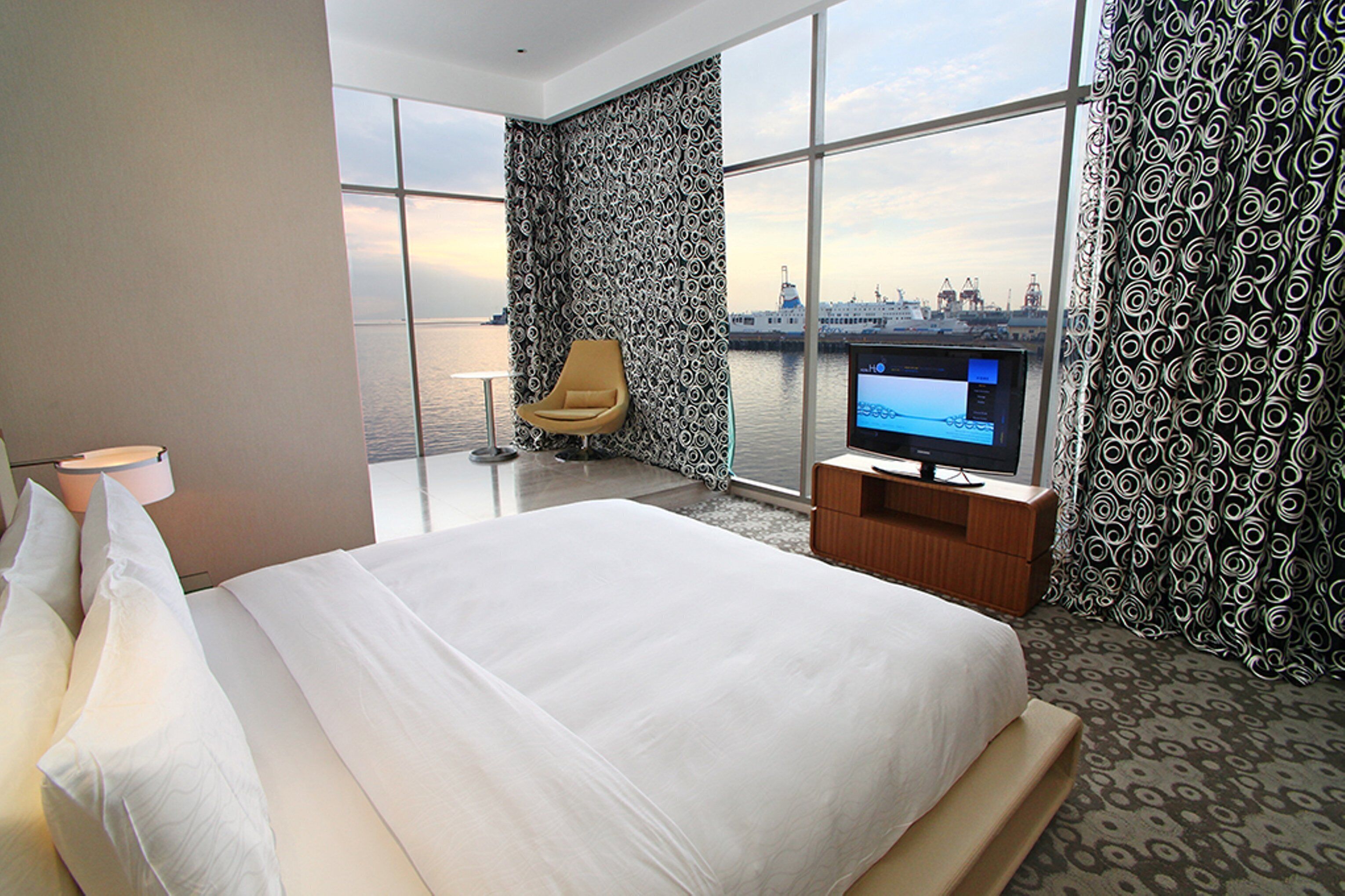 A hotel room with a large bed, a television, a chair, and a view of the water and city skyline.