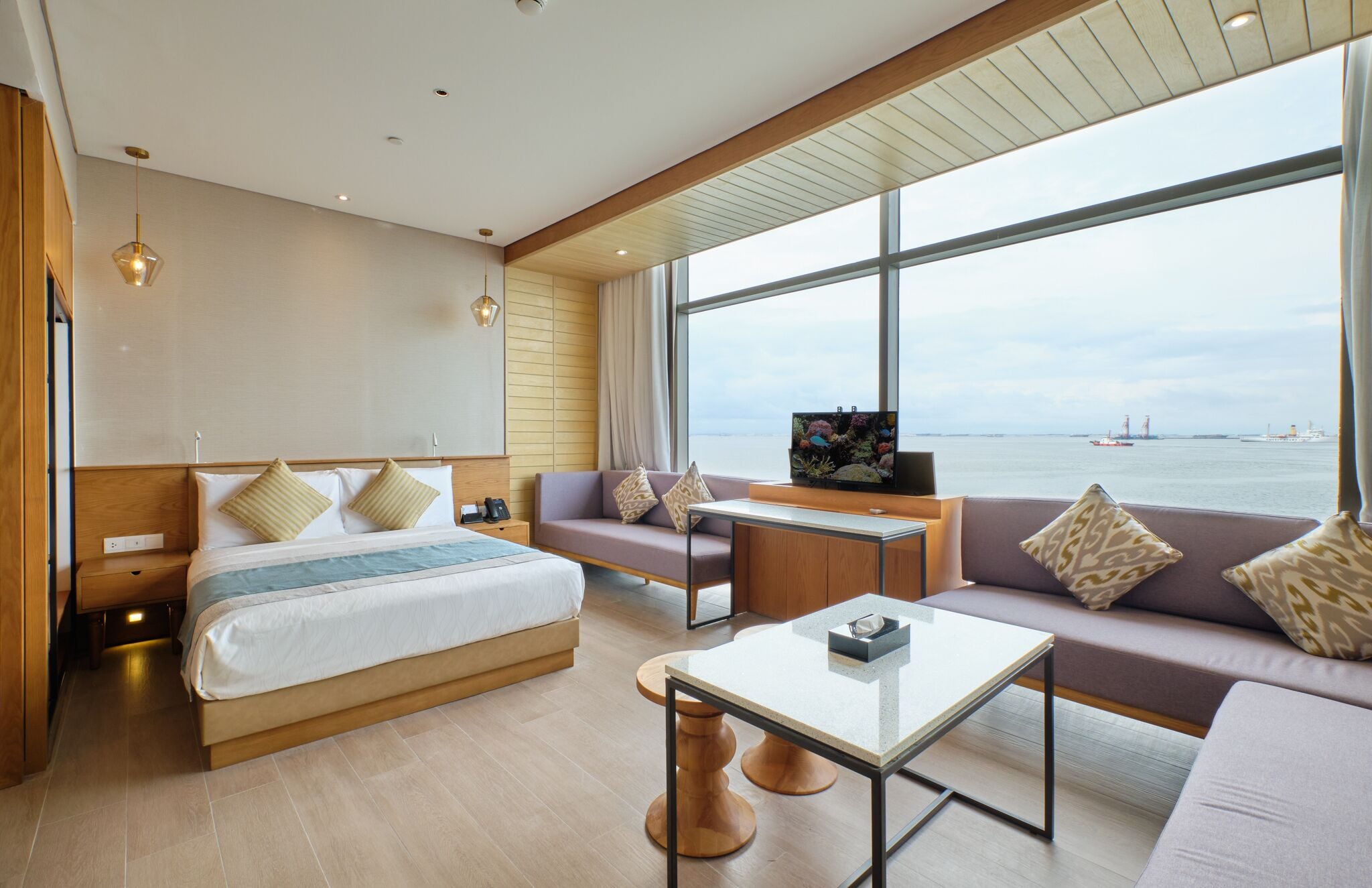 A modern hotel room with a large bed, a sofa, a small table, and a TV. The room has a large window with a view of the sea and ships.