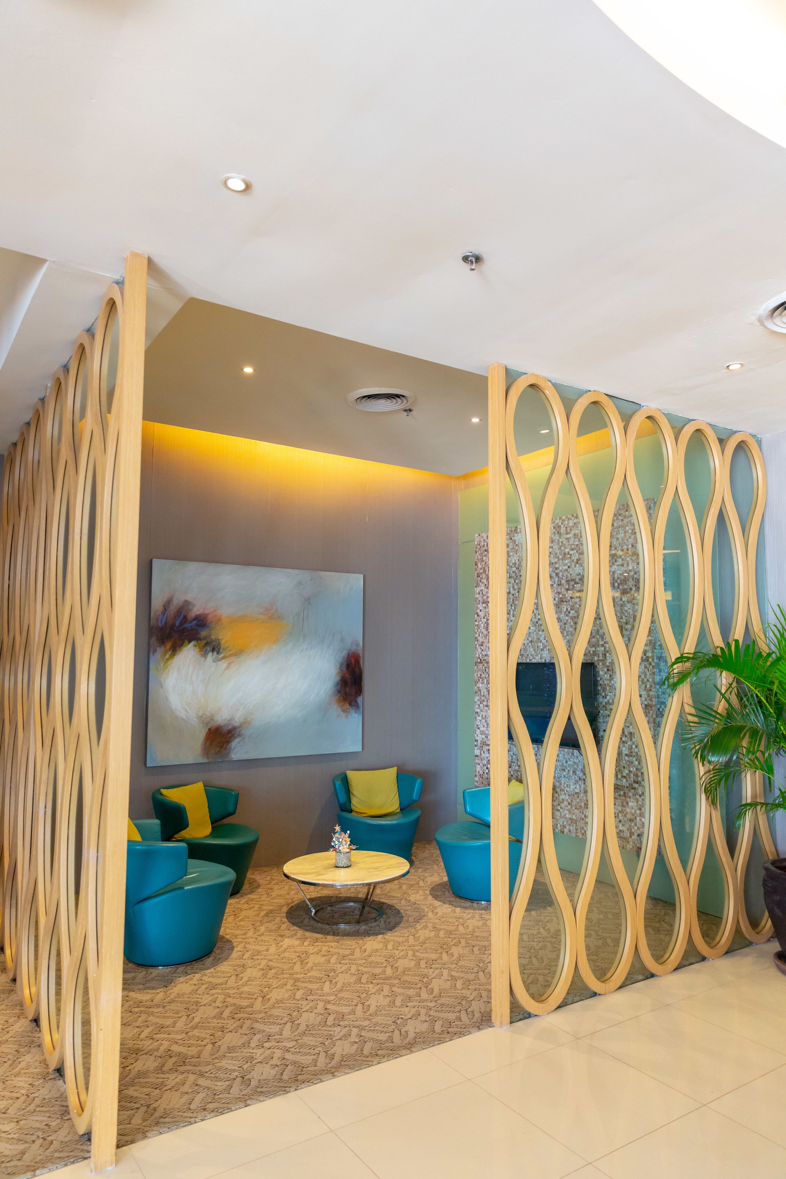 Un espace détente moderne agrémenté d’un grand tableau abstrait, de sièges aux couleurs turquoise et jaune, et d’une cloison décorative en bois.