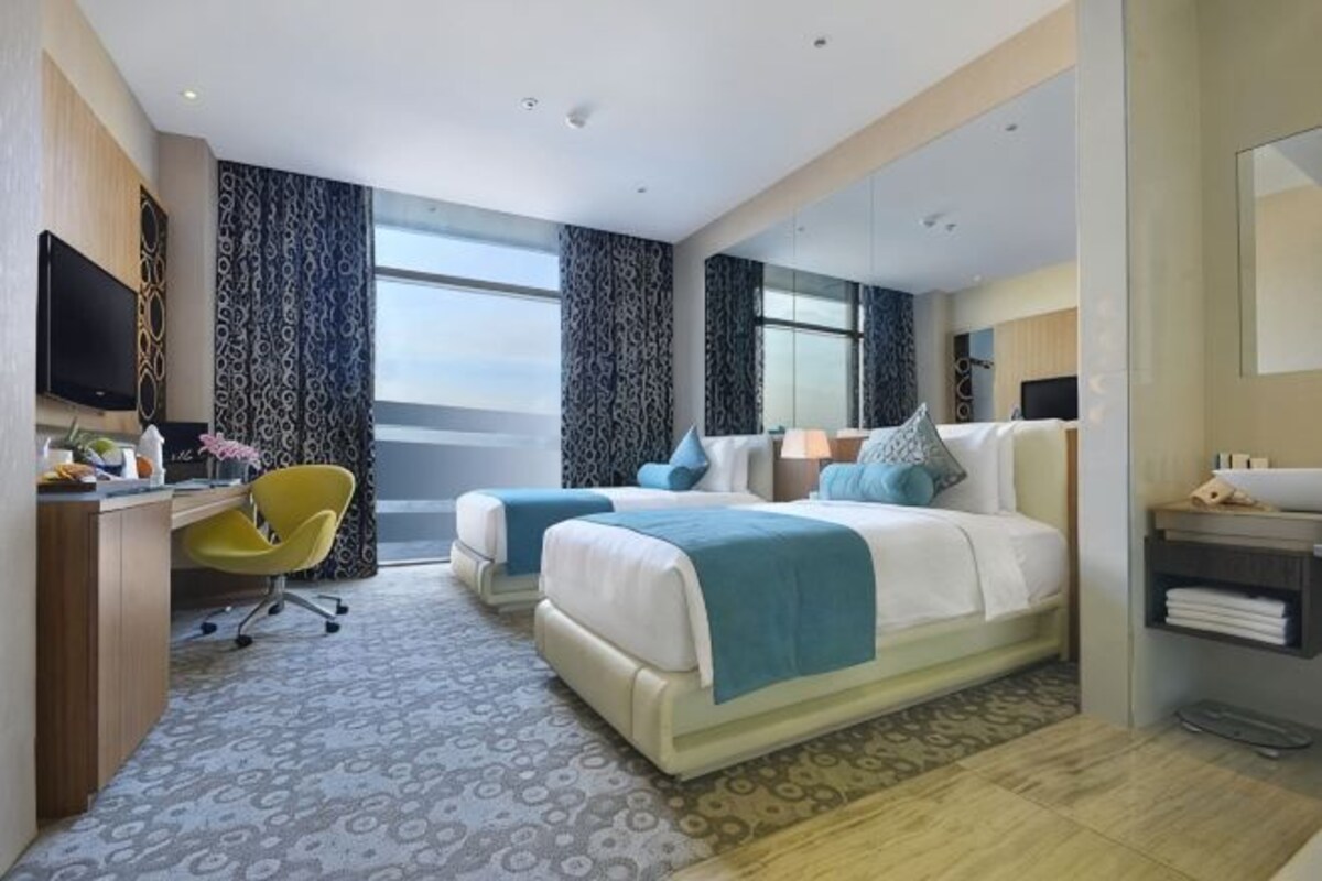 A hotel room with two beds, a desk with a chair, a television, and a large window.