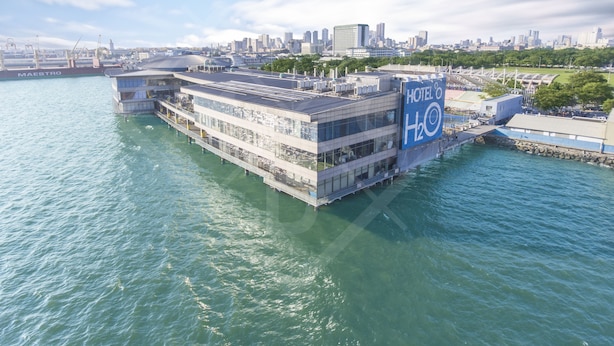 A waterfront hotel with a large glass facade, a prominent "HOTEL H2O" sign, and a view of the city skyline in the background. - Hotel H2O (Manila)
