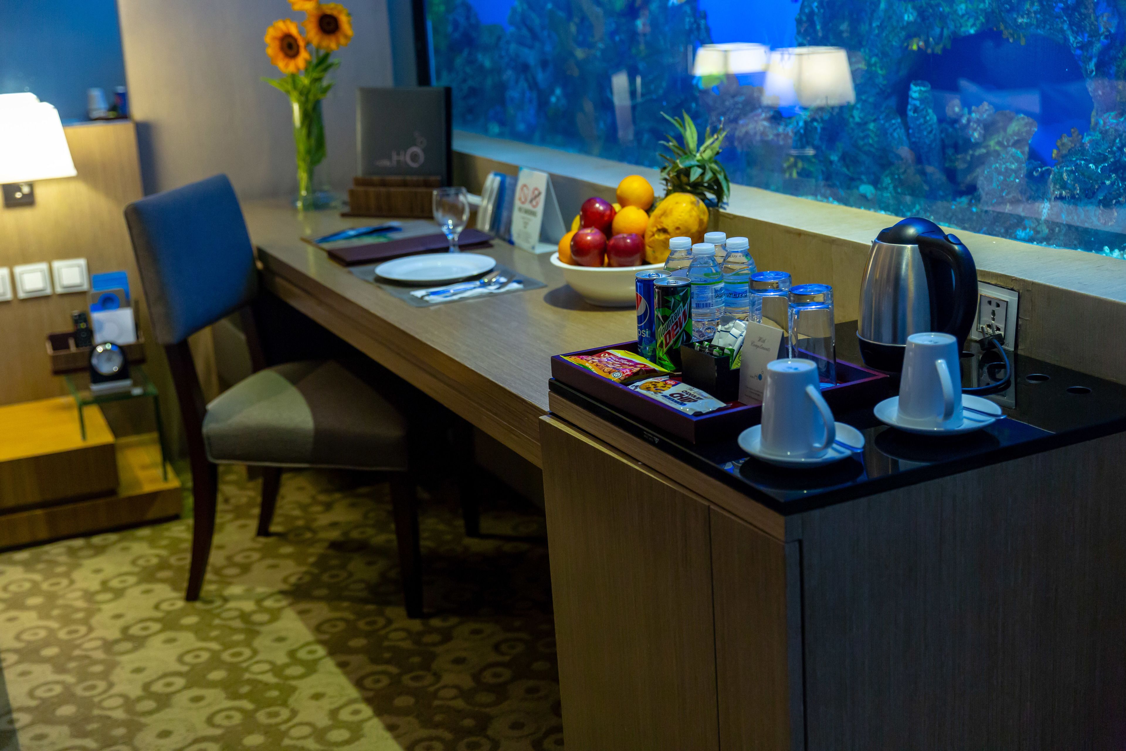 A hotel room with a dining area featuring a wooden table, a chair, a vase with sunflowers, a bowl of fruit, and a coffee maker.