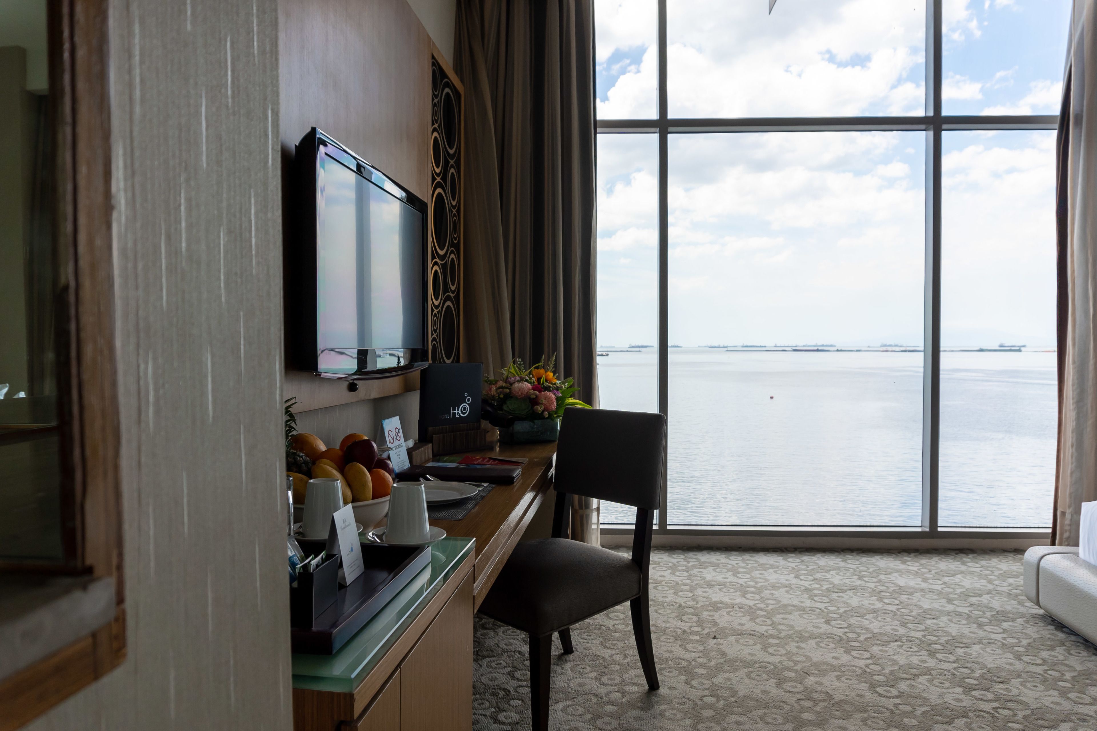 Une chambre d’hôtel dotée d’une grande fenêtre donnant sur la mer, d’un bureau avec une chaise, d’une télévision et d’une plante décorative.