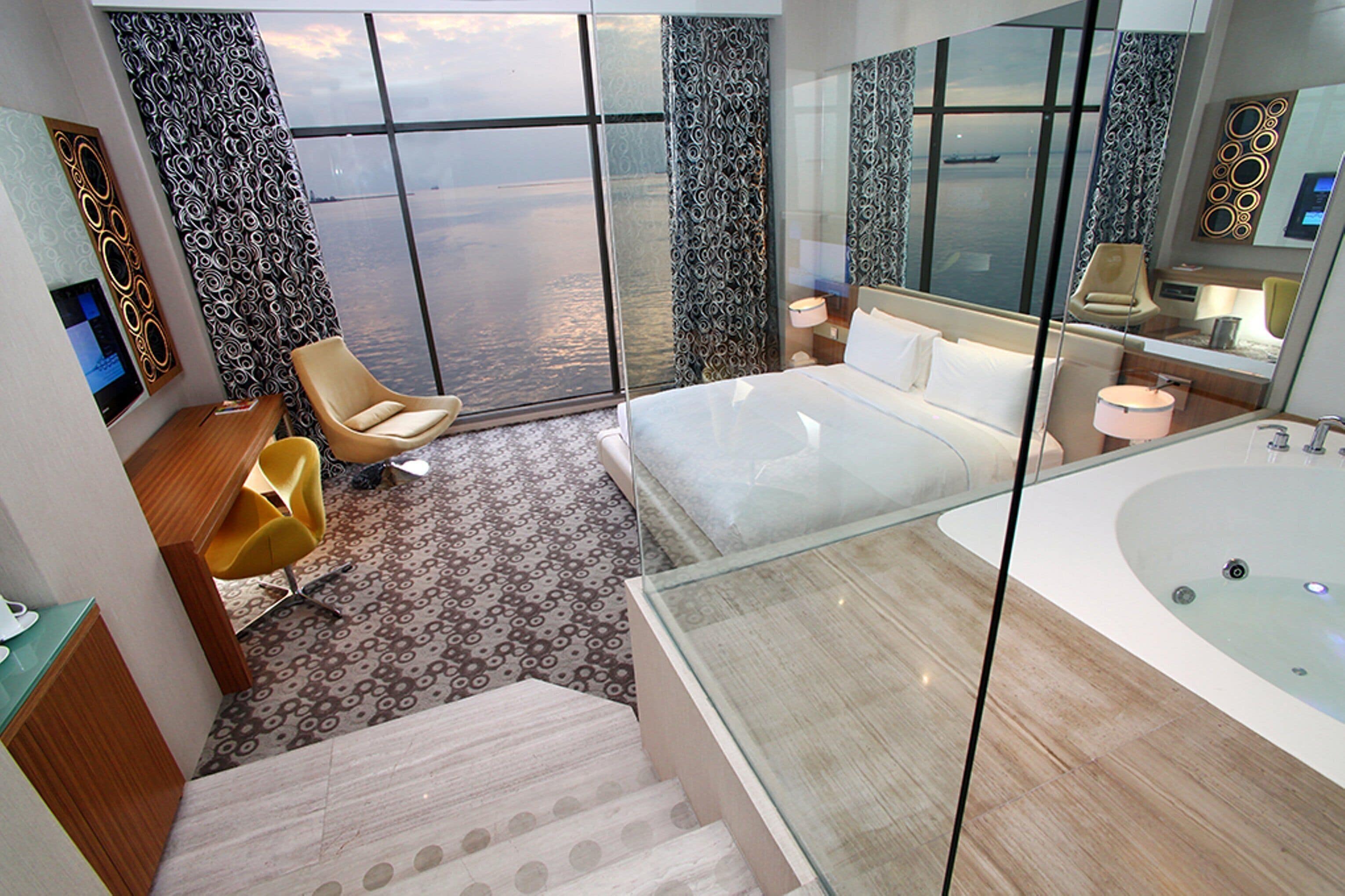 A modern hotel room with a large bed, a glass-enclosed bathtub, a seating area with a chair, and a view of the water.