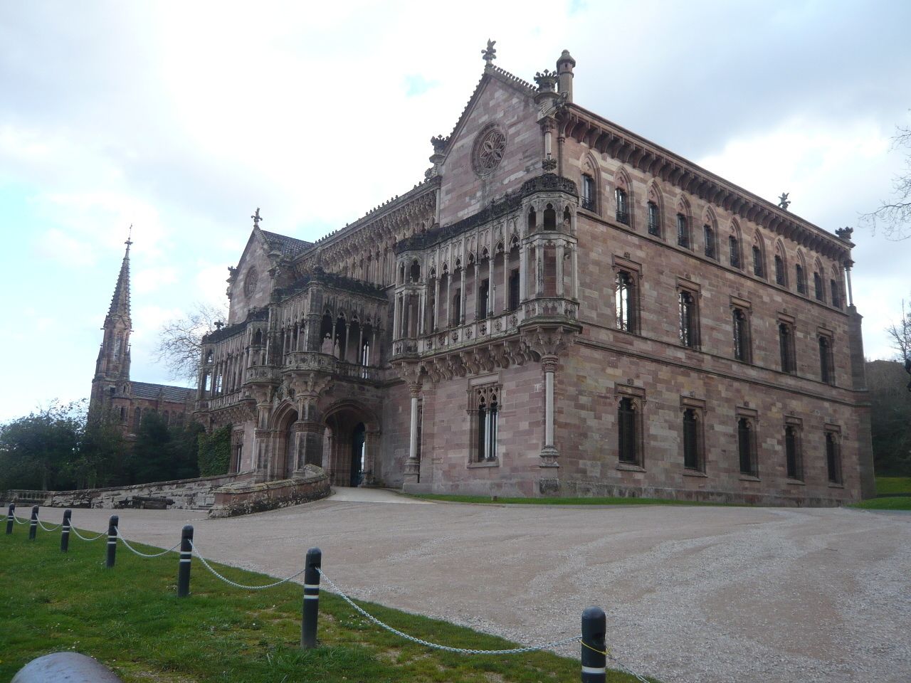 A historic building with a large arched entrance, a clock tower in the background, and a paved courtyard.