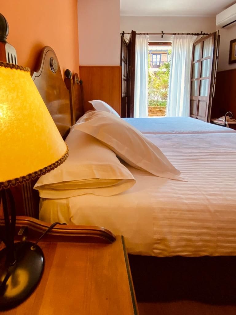 A hotel room with a bed, a desk with a lamp, and an open window.