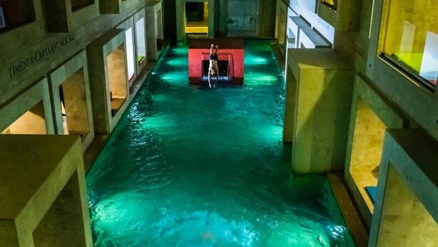 En indendørs swimmingpool med en springplatform i midten, omgivet af betonvægge med vinduer.