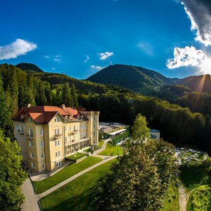 A large, multi-story building with a red roof, surrounded by lush greenery and mountains in the background. - Rimske terme - Hotel Sofijin Dvor (Lasko)
