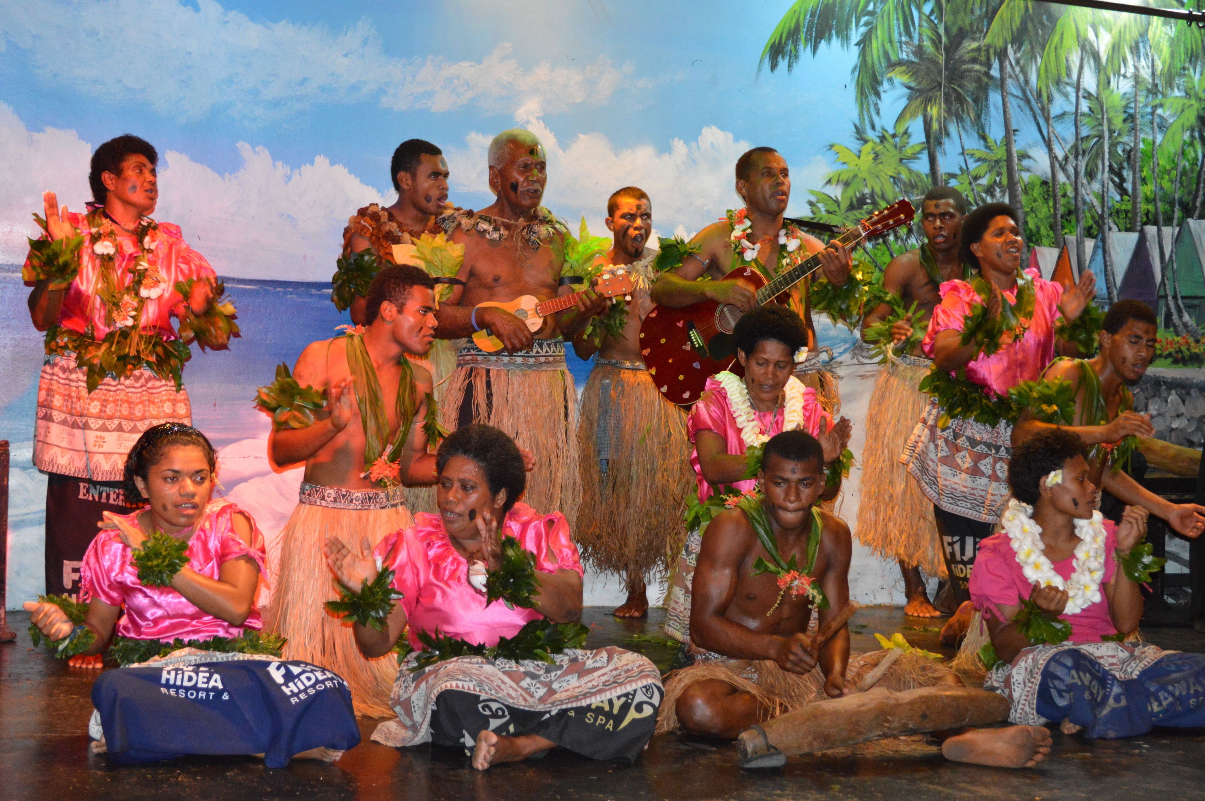 Eine Gruppe von Menschen in traditioneller Kleidung auf einer Bühne mit Palmen und einer Strandkulisse im Hintergrund.