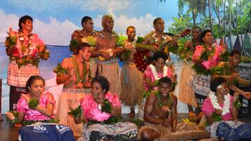 Eine Gruppe von Menschen in traditioneller Kleidung auf einer Bühne mit Palmen und einer Strandkulisse im Hintergrund.