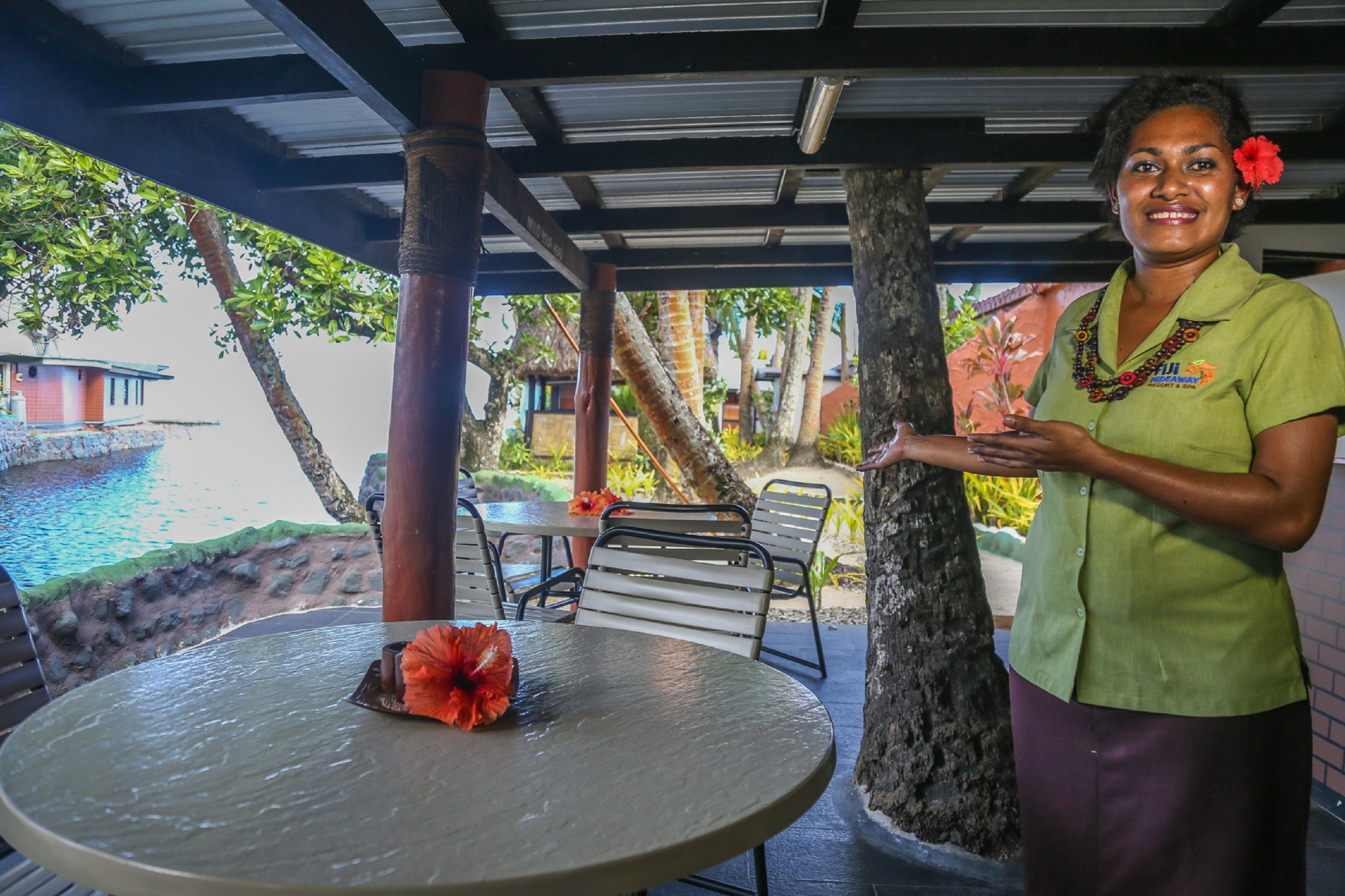 A covered outdoor seating area with a table and chairs, a tropical environment with trees and a view of a body of water.