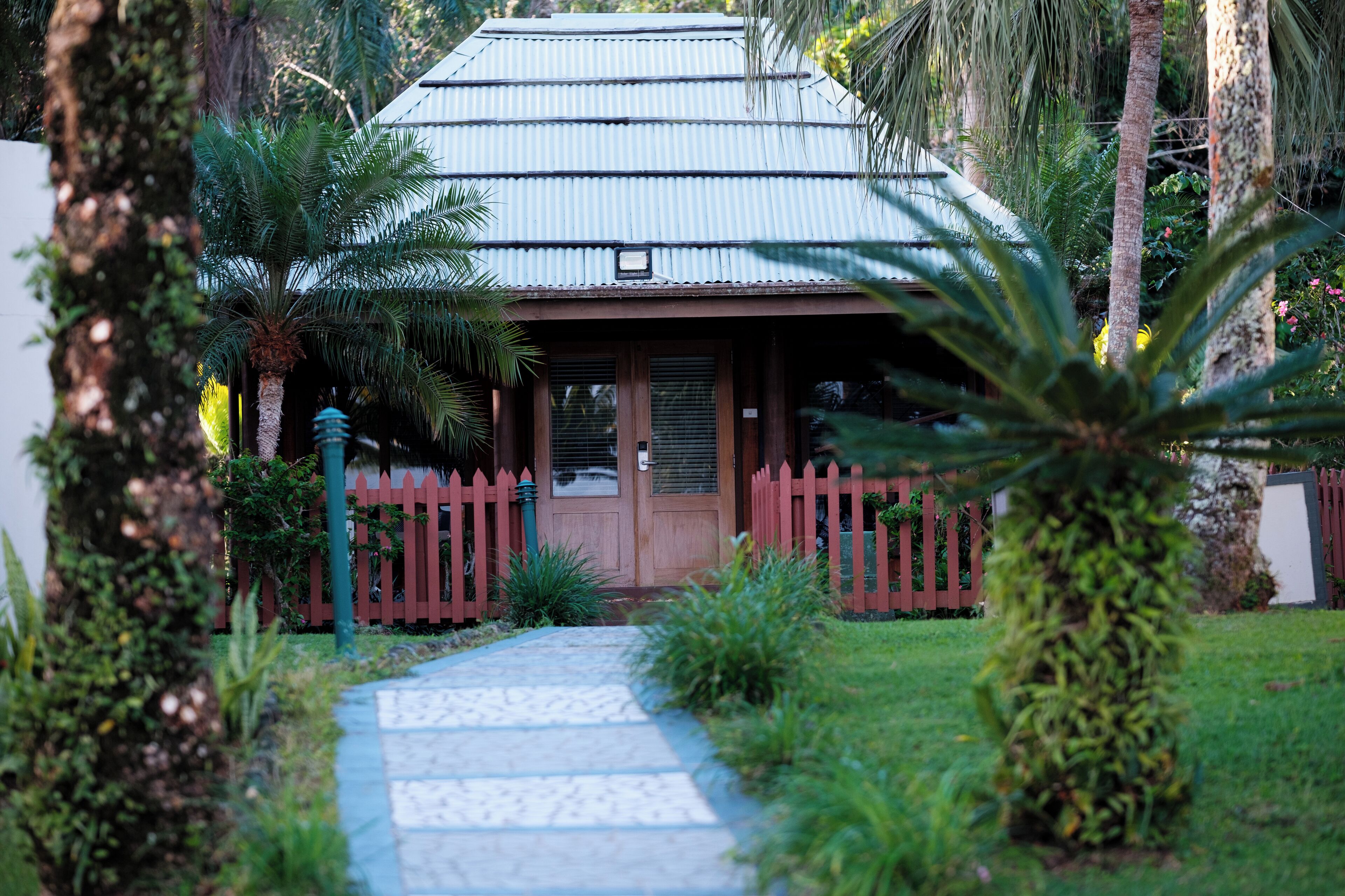 A house with a red picket fence, a pathway leading to the entrance, and tropical vegetation.