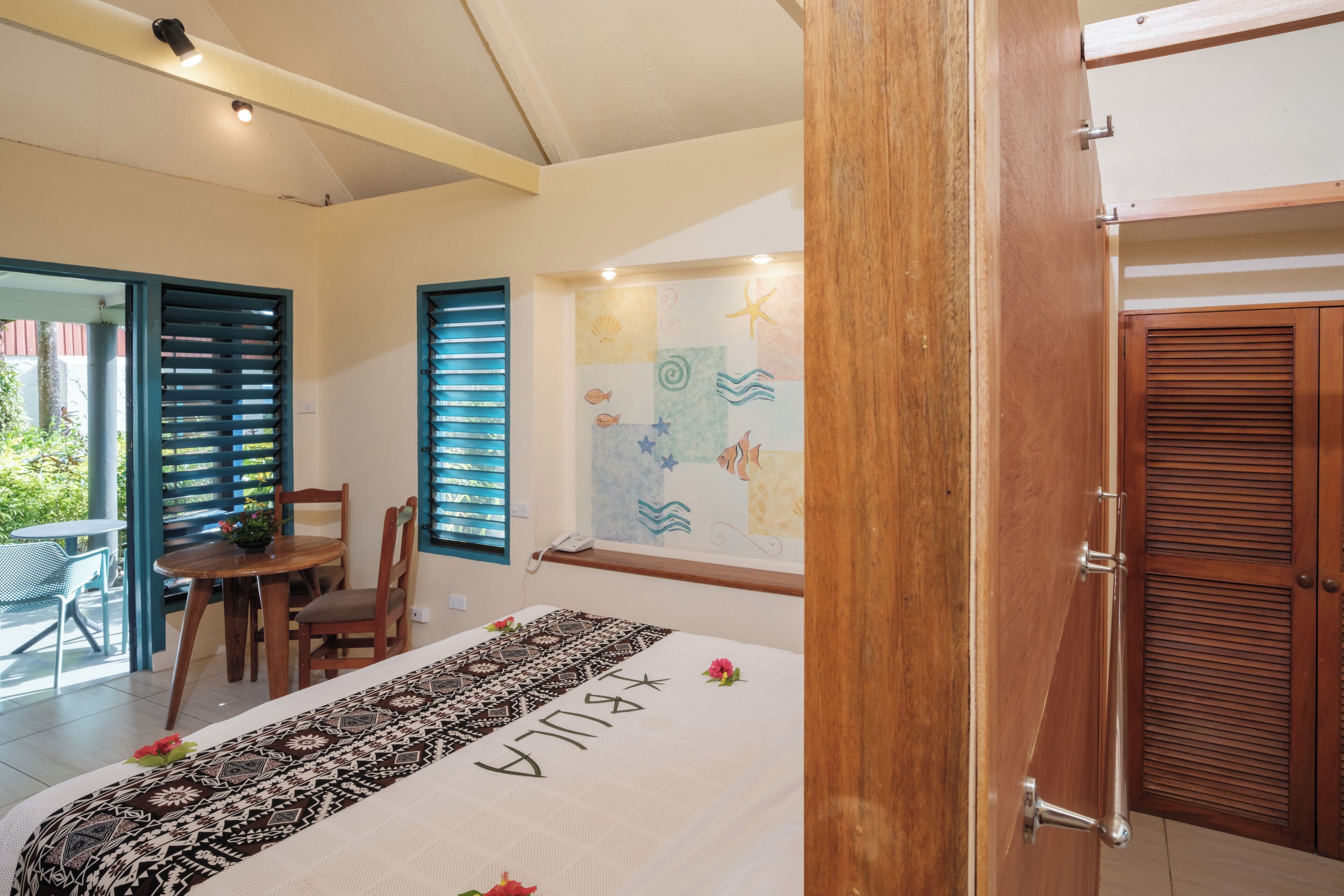 Ein Schlafzimmer mit einem Bett, einer Holztür, einem Fenster mit Jalousien, einem Wandbild mit Meeresmotiven und einem kleinen Tisch mit Stuhl.
