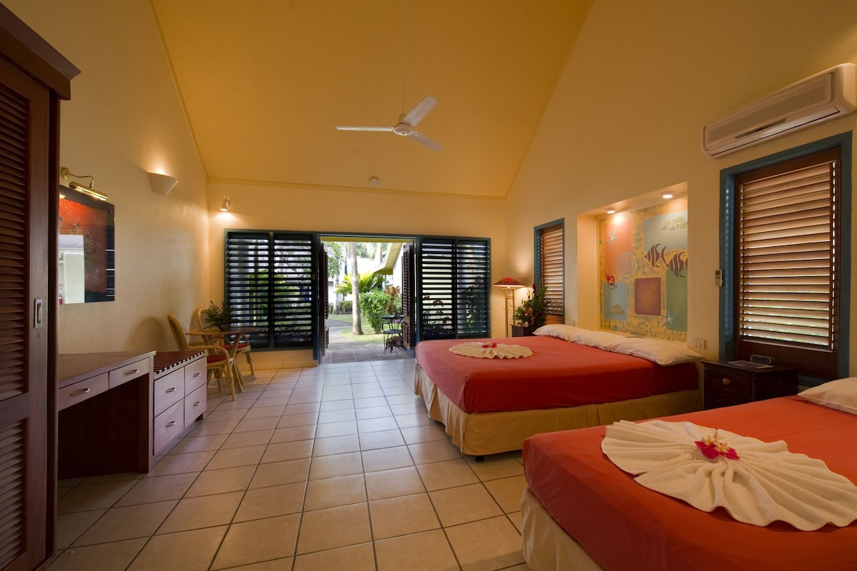 A hotel room with two beds, a desk, a chair, a ceiling fan, and a view of the garden.