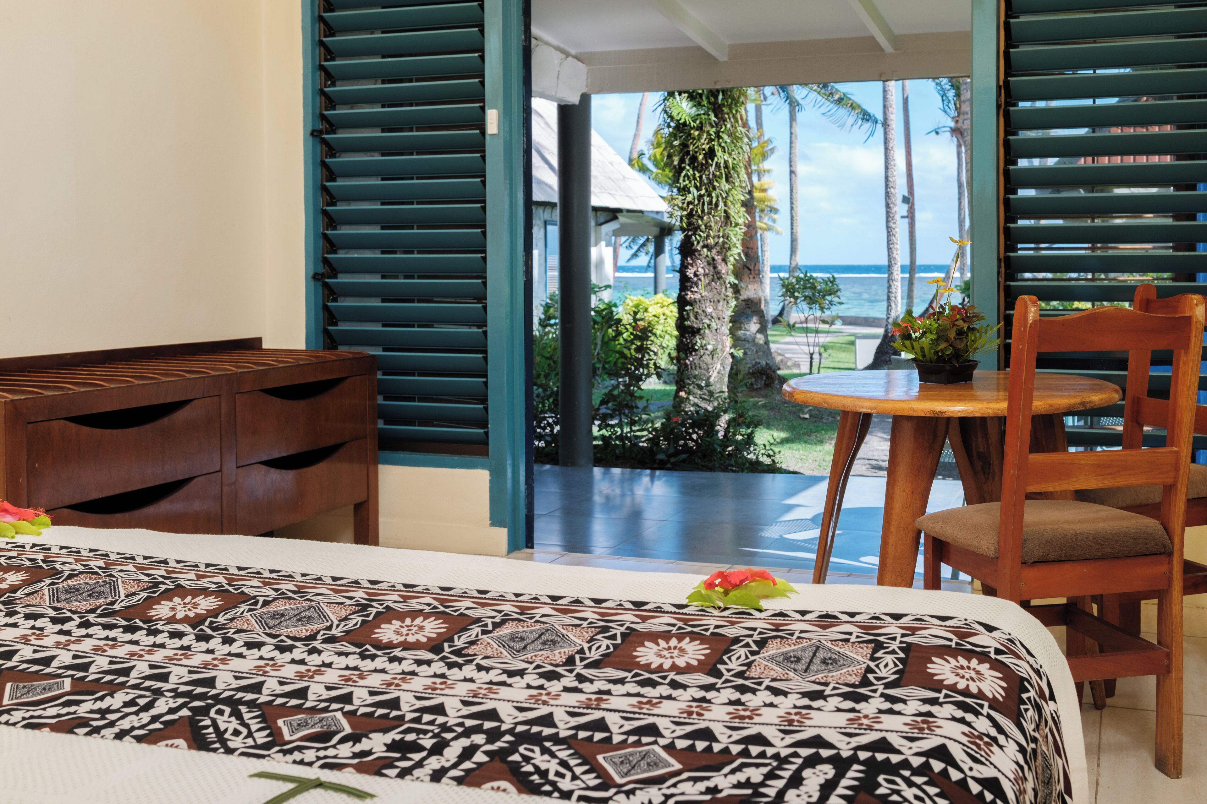 A hotel room with a bed, a wooden nightstand, a small table with a chair, and a view of the ocean through large windows.
