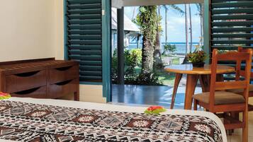 A hotel room with a bed, a wooden nightstand, a small table with a chair, and a view of the ocean through large windows.