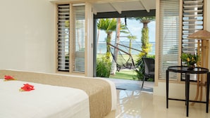 A bedroom with a bed, a small table with a lamp, and a view of the ocean through open shutters. - Fiji Hideaway Resort and Spa (Korolevu)