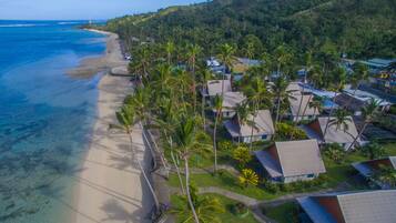 An aerial view of a coastal resort with beachfront villas, palm trees, and a clear view of the ocean.