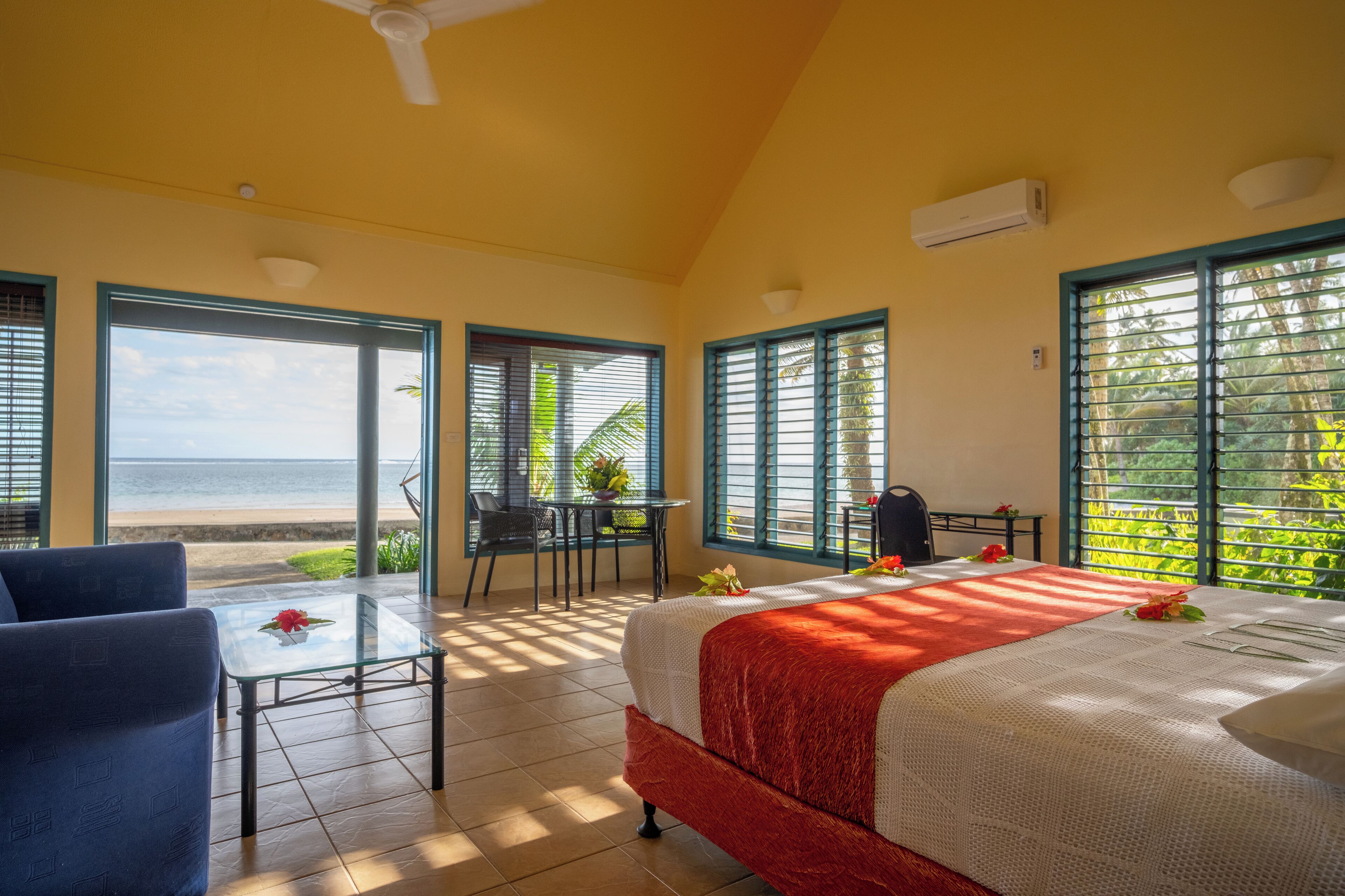 A room with a bed, a table, and a chair, with a view of the beach and palm trees.