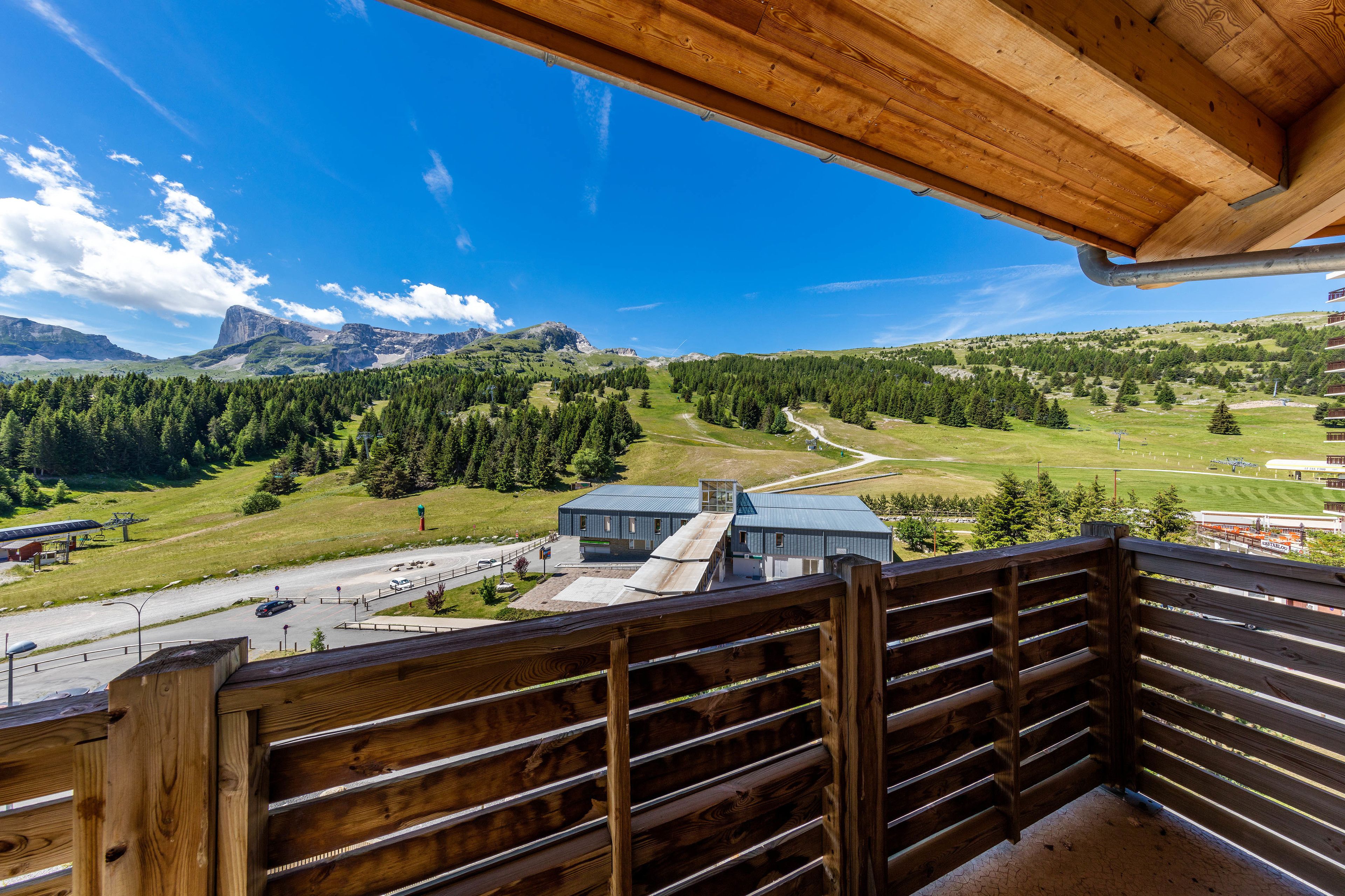 A wooden balcony with a view of a mountainous landscape, a road, and a building with a blue roof.