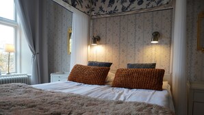A bedroom with a large bed, two wall-mounted lamps, a radiator, and a window with curtains. - Stjärnholms slott (Oxelosund)