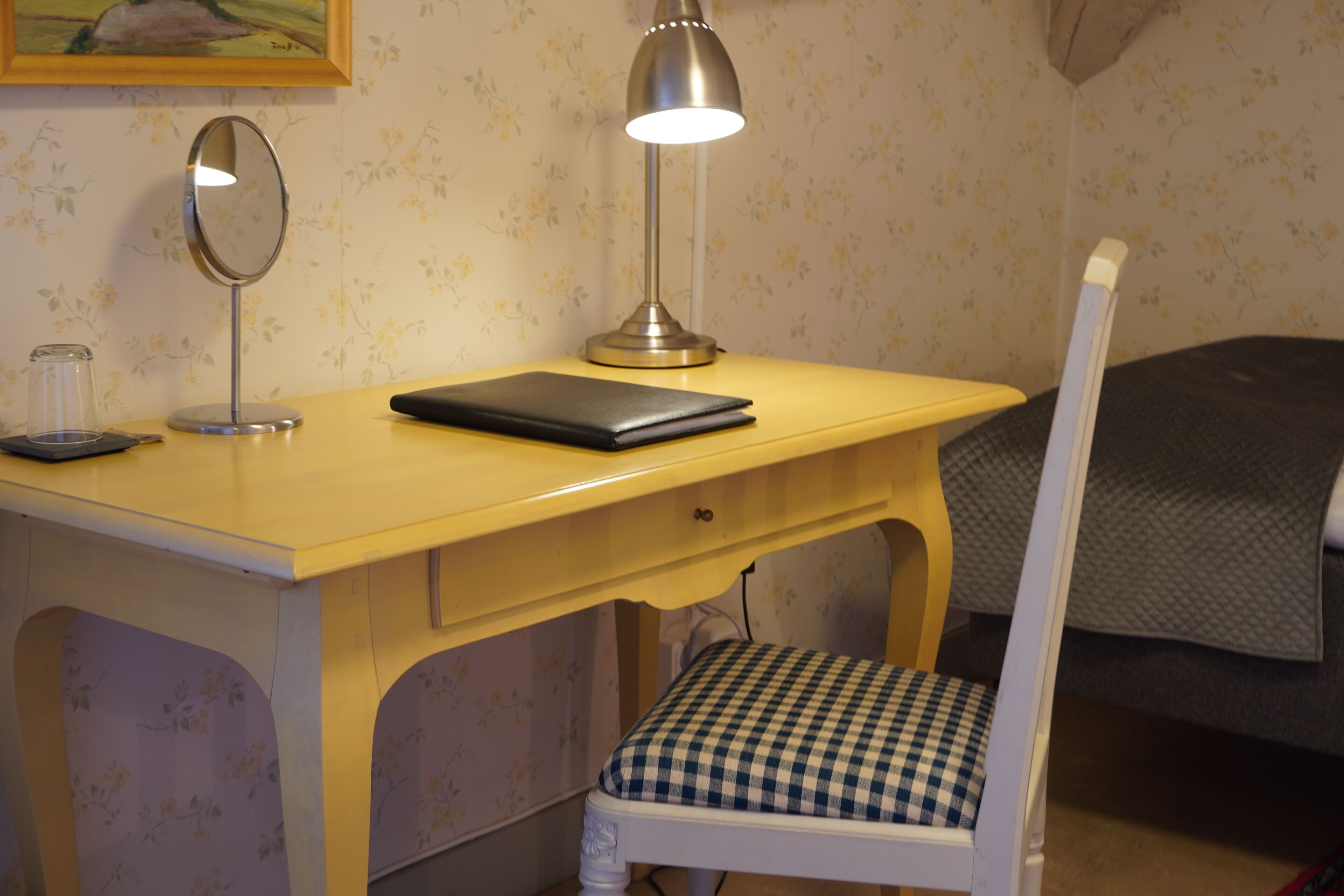 A small, neatly arranged bedroom with a single bed, a bedside table with a lamp, a chair, and a desk with a laptop.