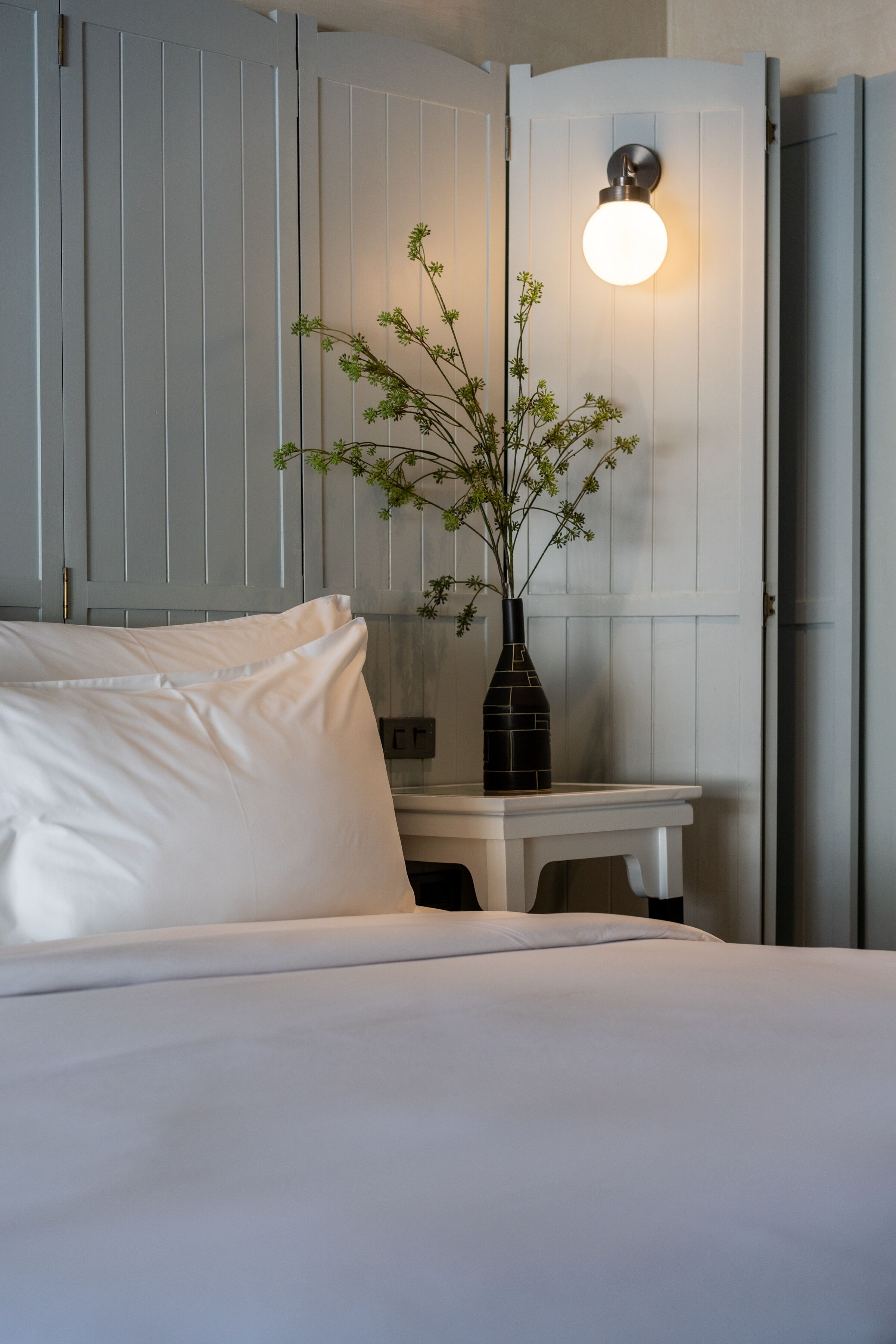 A neatly made bed with white linens, a small white table with a vase of green plants, and a wall-mounted light fixture.