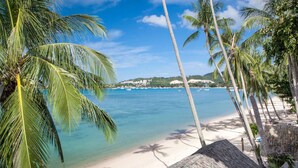A beach with palm trees, clear blue water, and a distant shoreline with buildings. - B House Samui (Koh Samui)