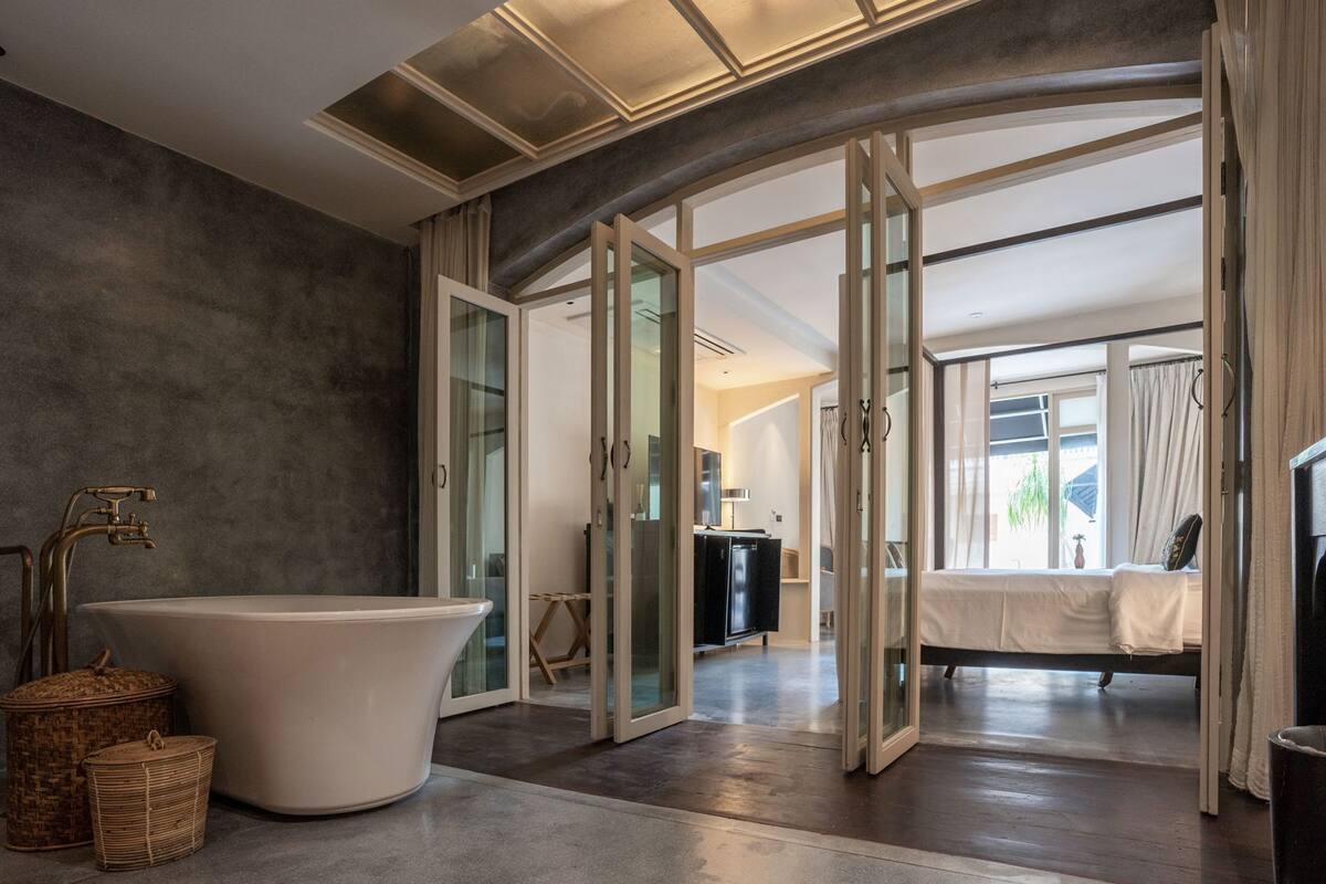 Un baño moderno con una bañera independiente, puertas de cristal que dan a un dormitorio y una cama a la vista.