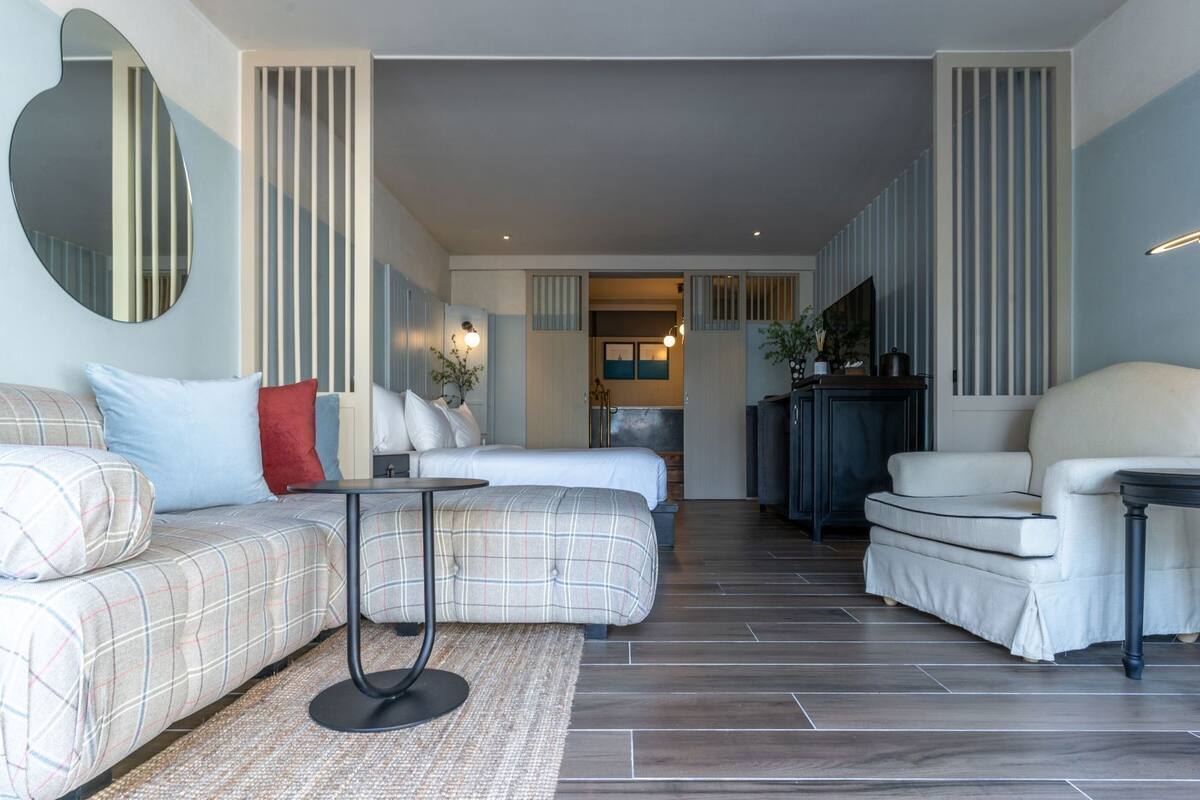 Habitación de hotel moderna con sofá, sillón y una pequeña mesa. Se ve una cama al fondo, un espejo en la pared y una vista al pasillo.