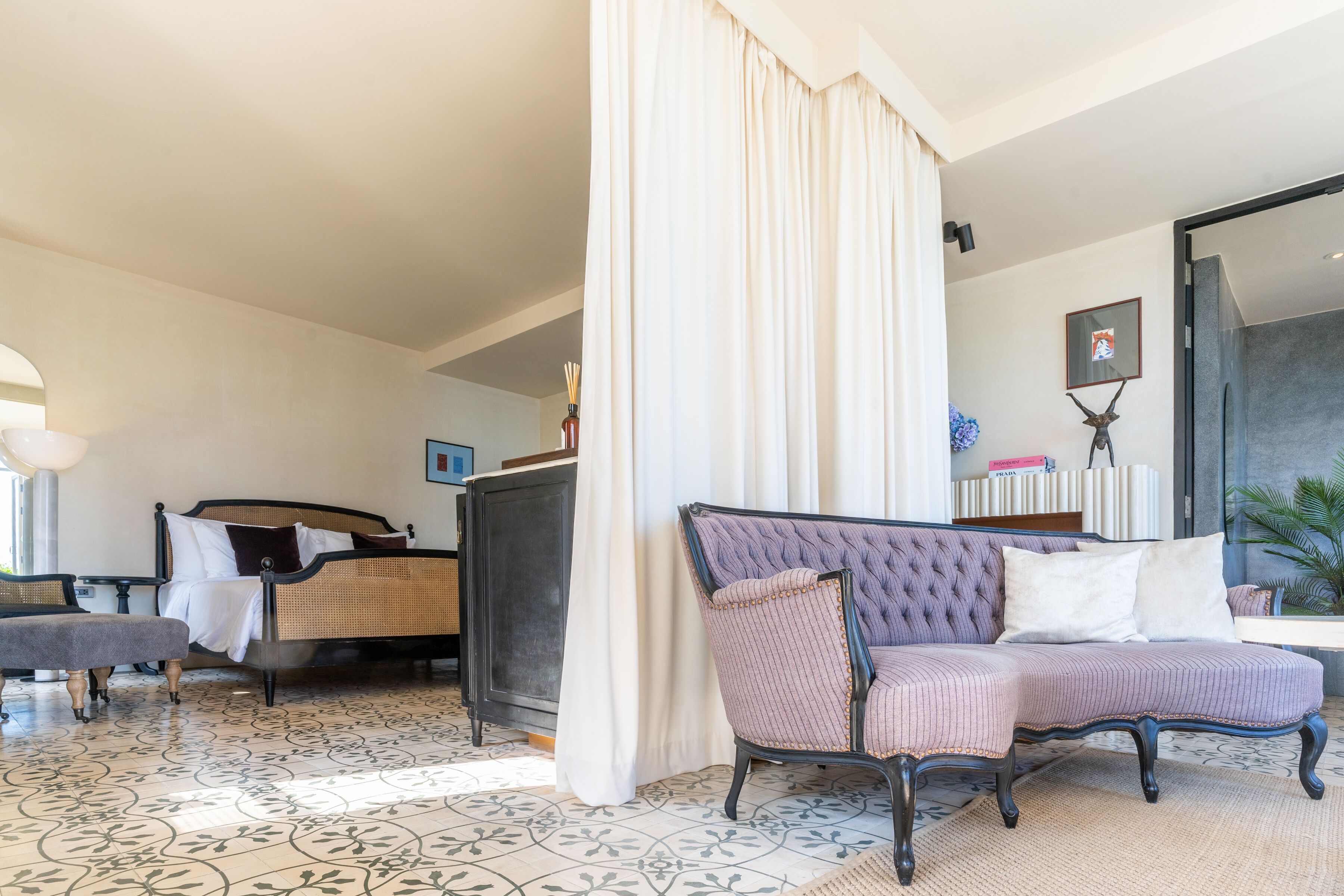 Un dormitorio con cama, una chaise longue, una mesita de noche y un ventanal con cortinas.