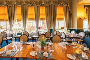 Ein Restaurant mit mehreren Esstischen, großen Fenstern mit Vorhängen und Blick auf Gebäude.