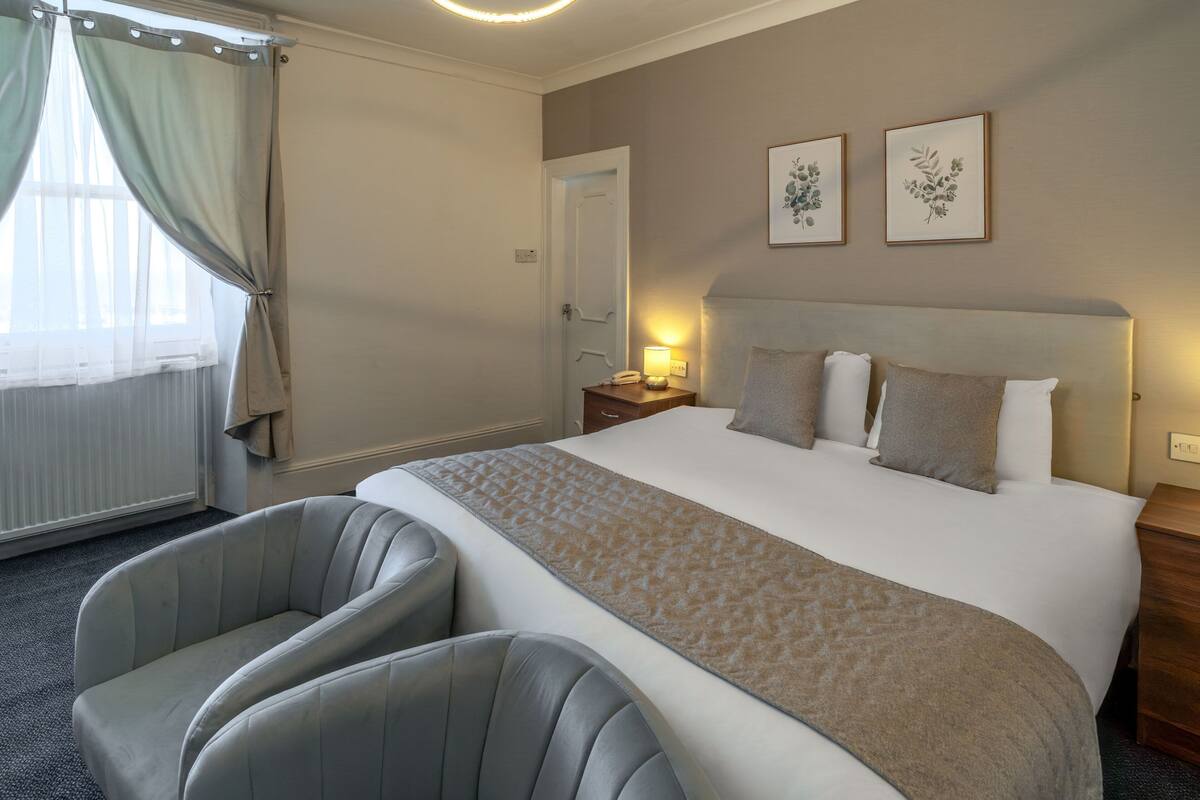 A hotel room with a bed, two pillows, a bedside table with a lamp, a chair, and a window with curtains.