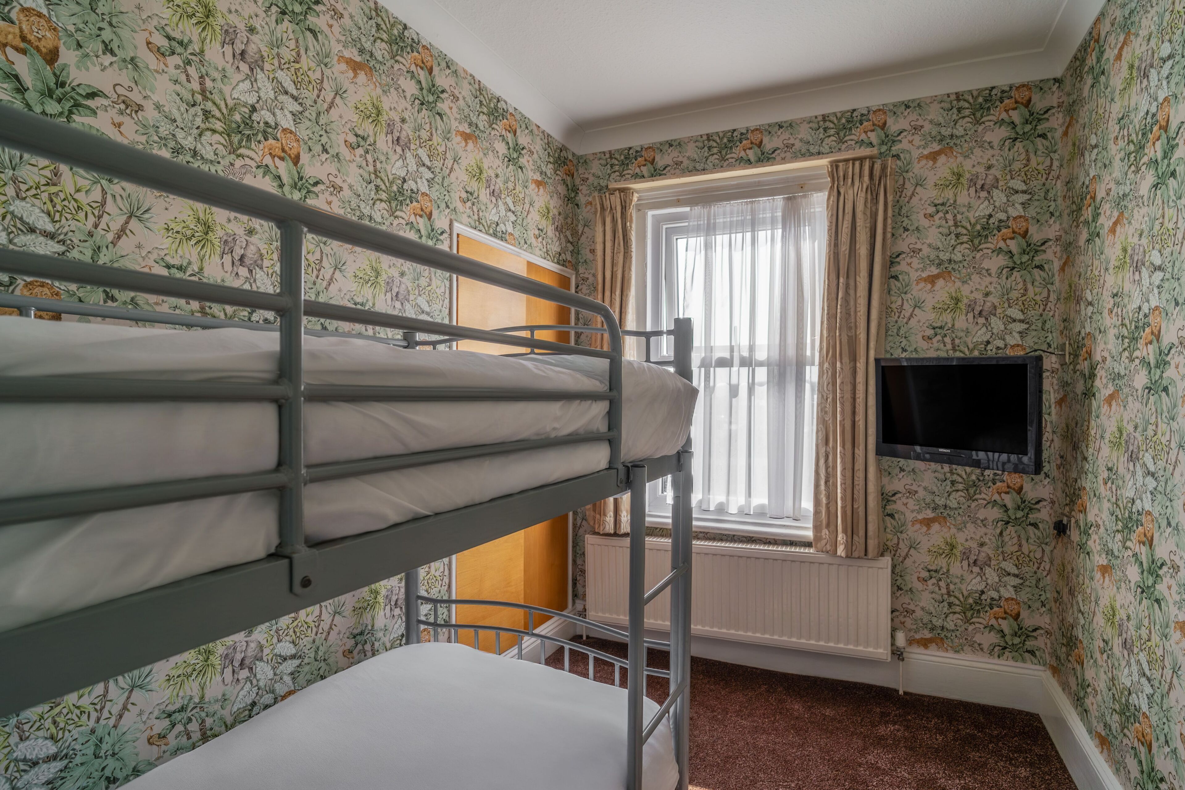 A bunk bed room with floral wallpaper, a window with curtains, and a TV mounted on the wall.