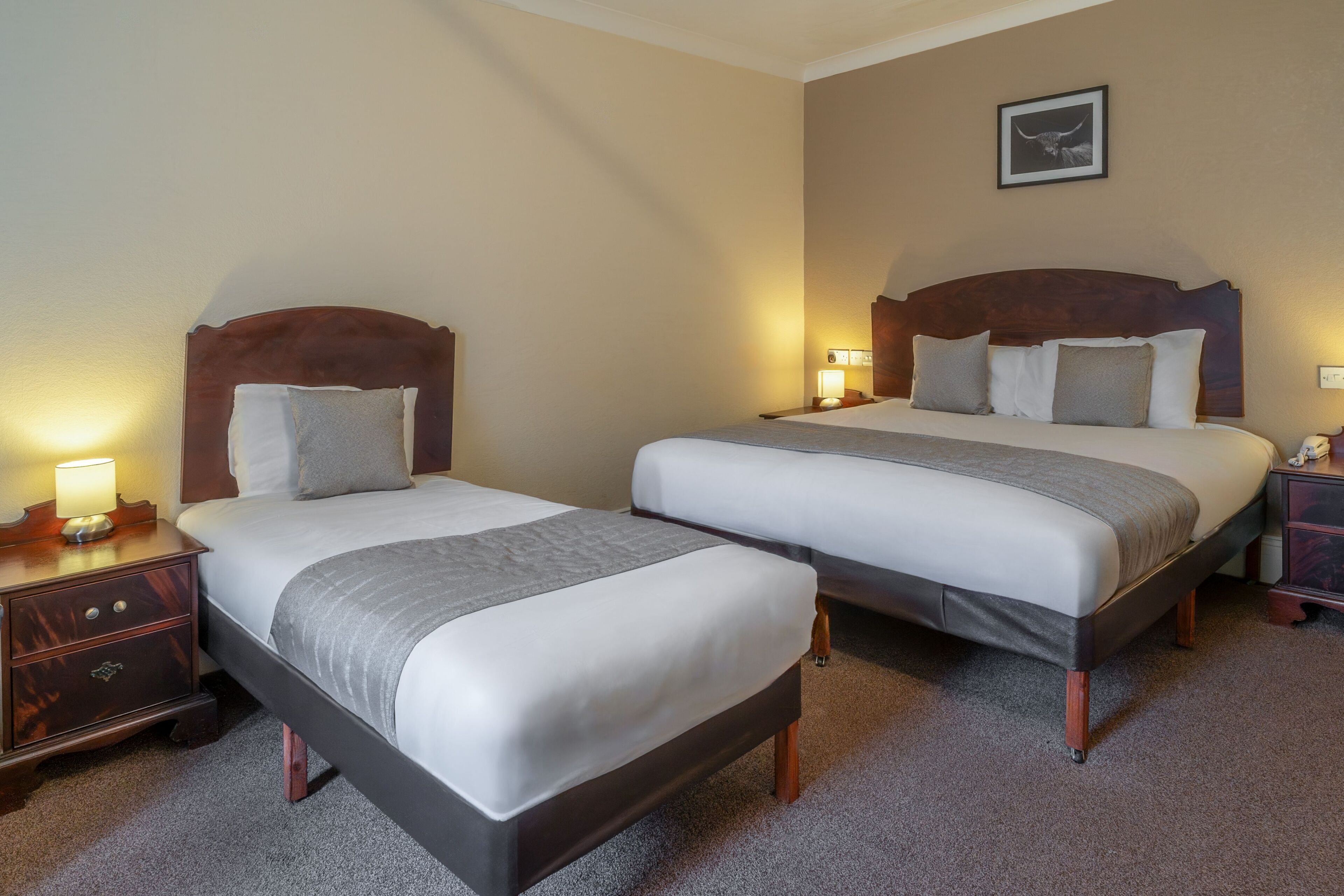 A hotel room with two beds, wooden headboards, bedside lamps, a nightstand, and a framed picture on the wall.