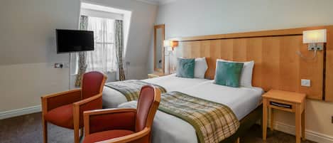 Habitación de hotel con dos camas, televisión, silla, mesita de noche y ventana con cortinas.