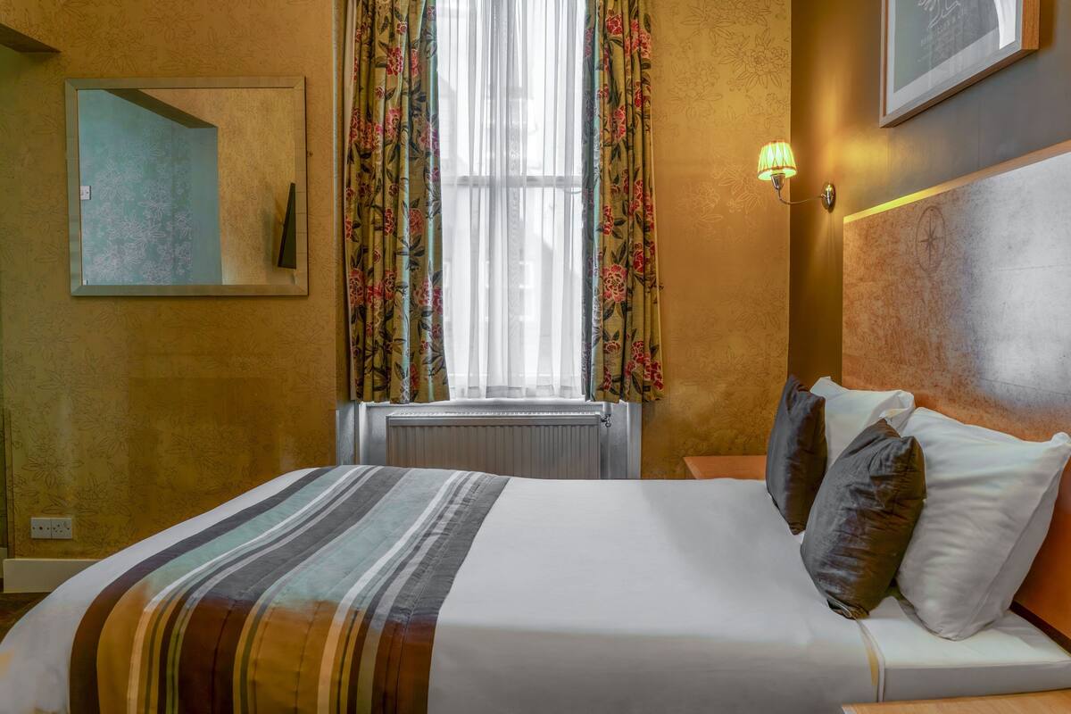 A hotel room with a bed, a window with floral curtains, a wall-mounted lamp, and a framed picture.