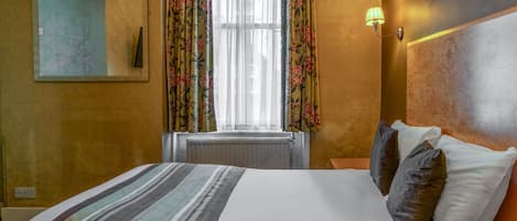 A hotel room with a bed, a window with floral curtains, a wall-mounted lamp, and a framed picture.