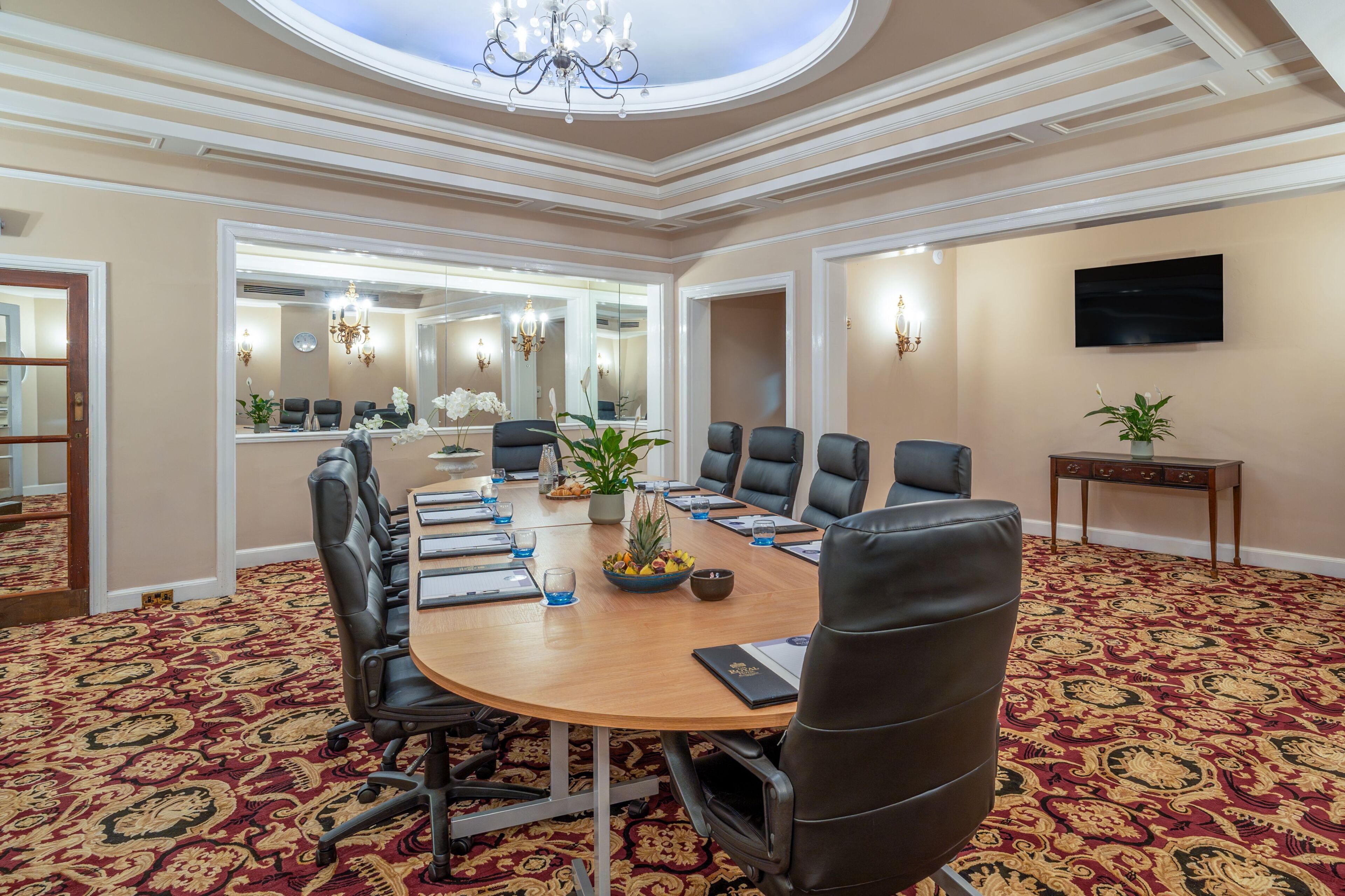 Une salle de conférence équipée d’une grande table, de chaises, d’un lustre, d’un téléviseur et d’un tapis à motifs.
