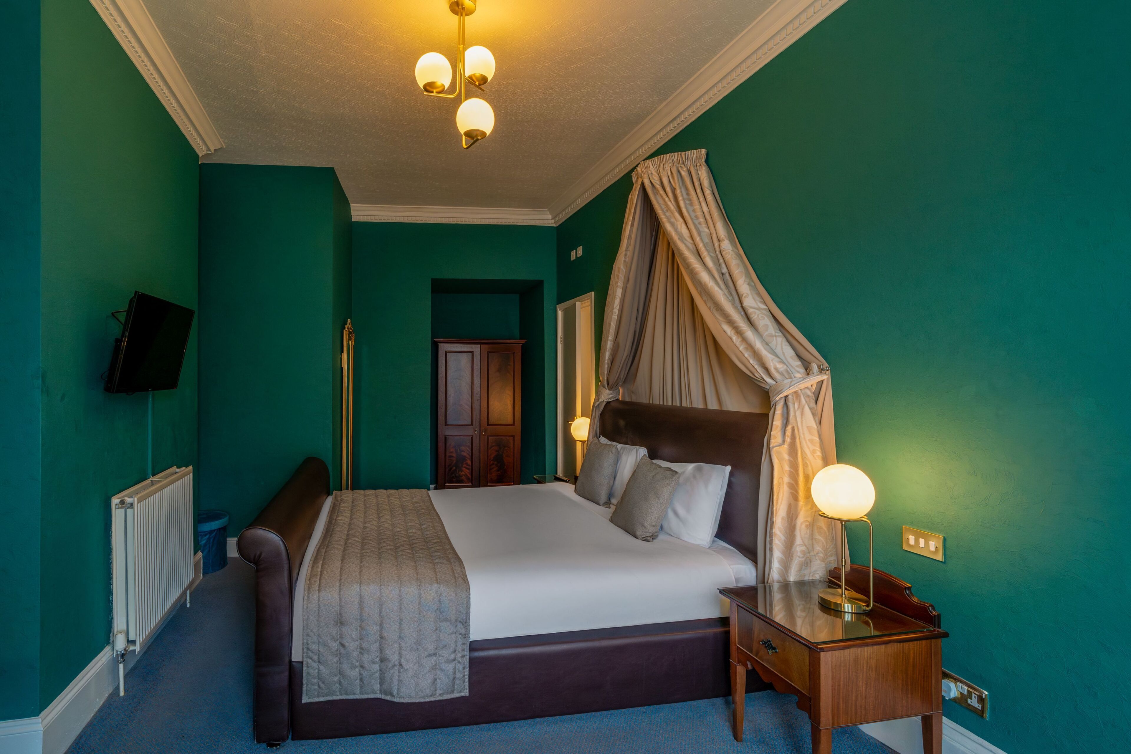 A bedroom with a large bed, a bedside table with a lamp, a radiator, and a television on the wall.