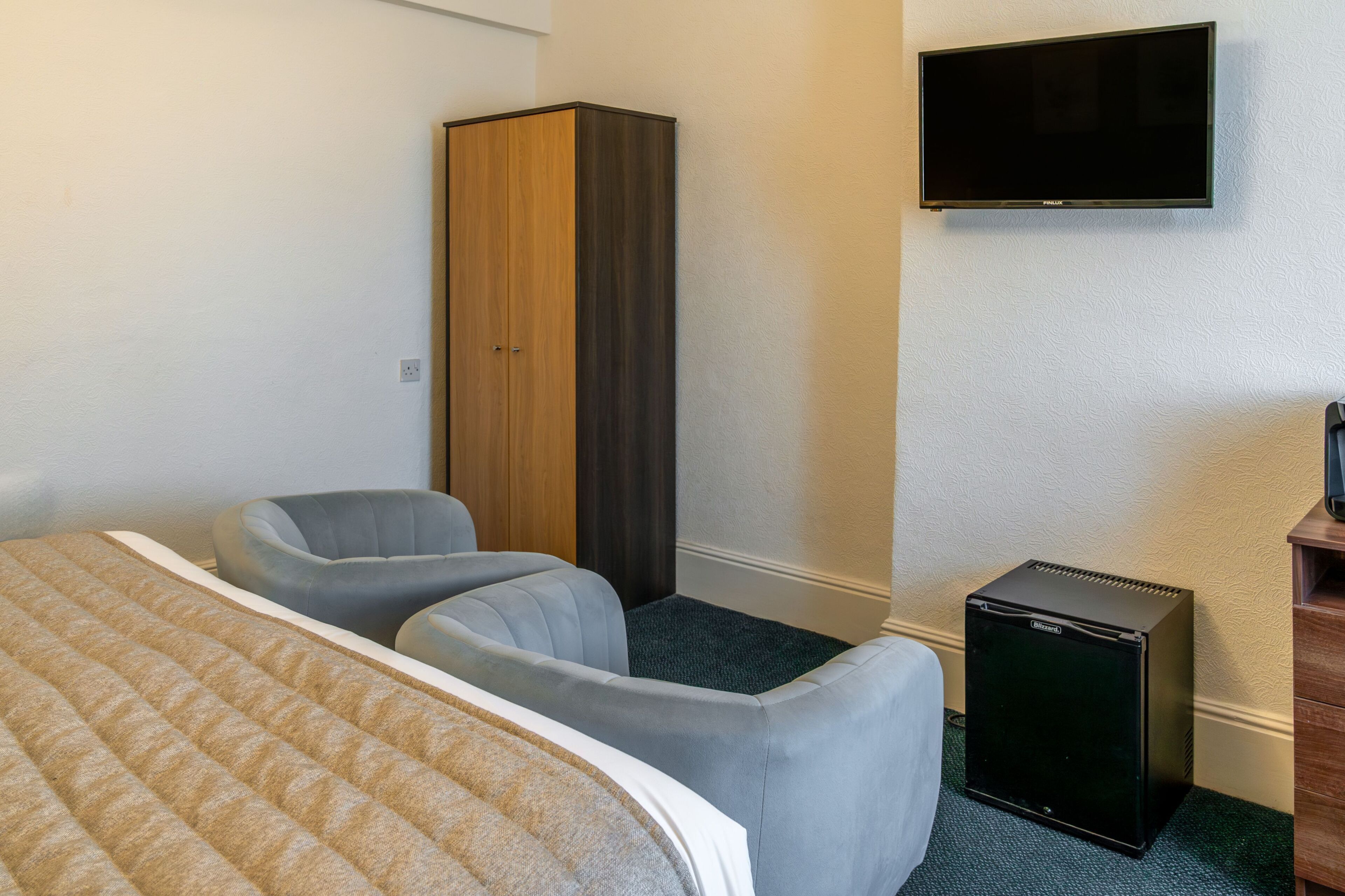 A hotel room with a bed, two armchairs, a wardrobe, a television, and a small refrigerator.